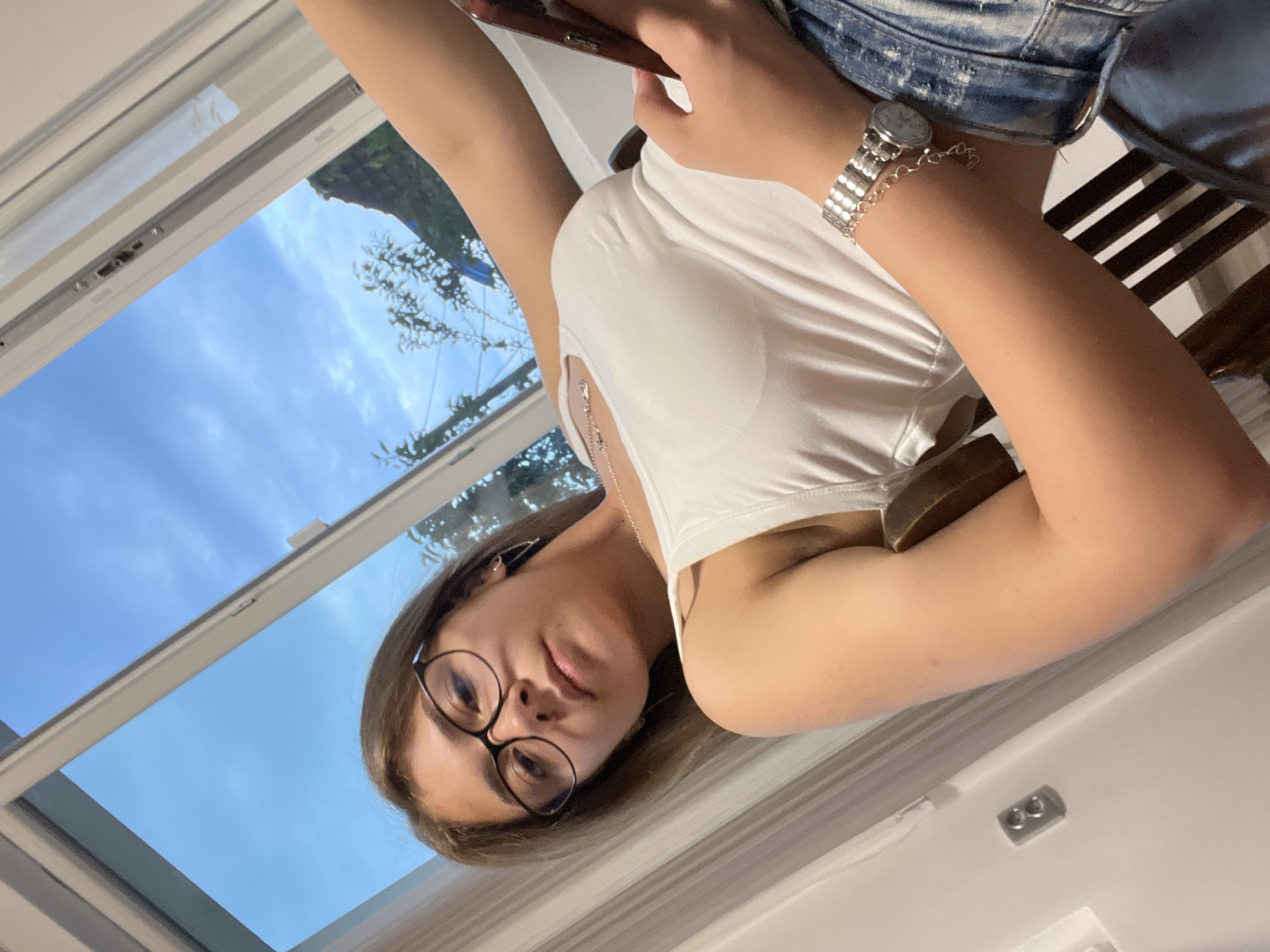 The image depicts a young woman seated indoors, looking directly at the camera, reportedly in Karavelovo, Bulgaria. She is positioned in a slight upward-angled shot, leaning against what appears to be a dark wooden chair with vertical slats, partially visible behind her right arm. The woman has brown hair, wears black-framed glasses, and a white sleeveless tank top. A silver-toned necklace with a small pendant is visible, along with mismatched earrings: a small stud on her right ear (viewer's left) and a longer, chain-link earring on her left ear (viewer's right). Her left wrist (viewer's right) is adorned with a silver-toned metal watch featuring a circular face and an attached charm. Her lower body, visible in the bottom right, shows blue denim jeans with some distressed detailing. Her left hand (viewer's right) holds a smartphone encased in a reddish-brown cover. To her left, a light-colored wall features a double electrical outlet. A large white-framed window dominates the upper and right portions of the background, revealing a clear blue sky with scattered white clouds and green foliage from trees or bushes outside. The window frame appears slightly askew or open at the top.