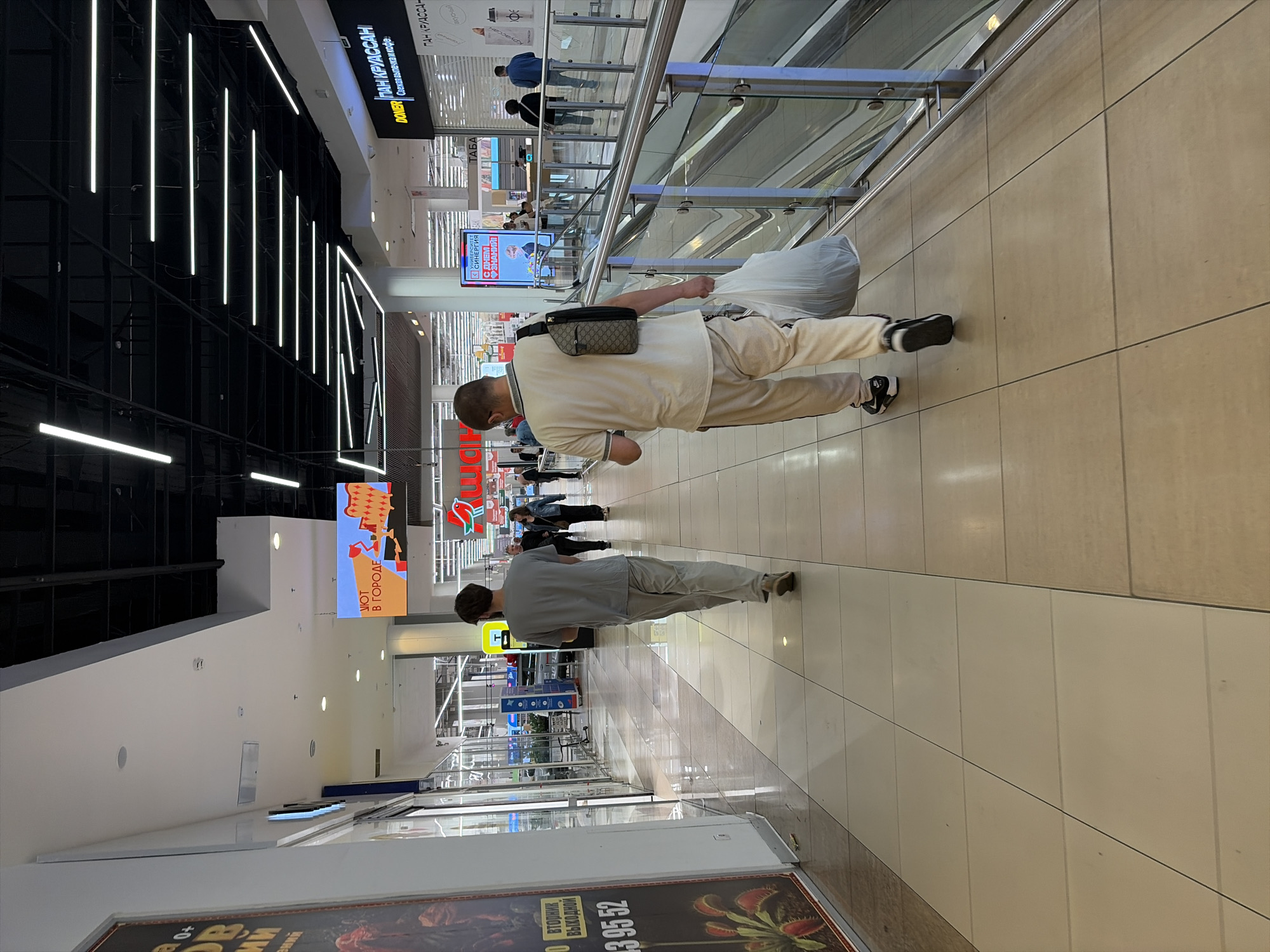 The image shows an interior view of a modern shopping mall, likely in Moscow, Russia, based on the Cyrillic text visible on signage. The scene is well-lit with overhead fluorescent and LED lighting. The floor is tiled in a light beige and brown pattern.
In the foreground, two men are walking away from the camera down a wide corridor. The man on the left is wearing a grey t-shirt and light-colored trousers. The man on the right is wearing a cream-colored polo shirt, matching light-colored track pants, and black athletic shoes. He is carrying a large white plastic bag in his right hand and has a dark patterned sling bag slung over his left shoulder.
The mall environment features glass storefronts on the left and a metal and glass railing on the right, overlooking a lower level or escalator. In the background, other shoppers are visible, as well as various store signs and digital displays. One digital display shows an animated advertisement with Cyrillic text. Another prominent sign reads "DONER ПАН КРУАССАН" with additional text below in Russian, indicating food establishments. The overall impression is one of a bustling, contemporary retail space.