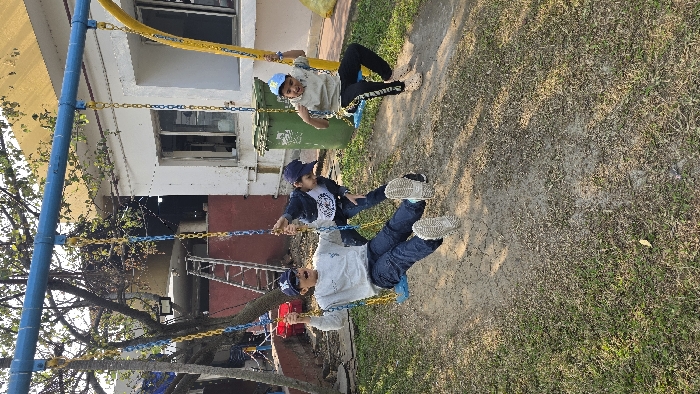 Two children play on swings in a playground. One child wears a beige shirt, dark pants, and a blue cap, while the other wears a white shirt, denim jacket, jeans, and a blue cap. They are positioned at different heights on the swings. A building is visible in the background, along with a yellow slide, a green trash can, and various trees. The ground is covered in grass and dirt.