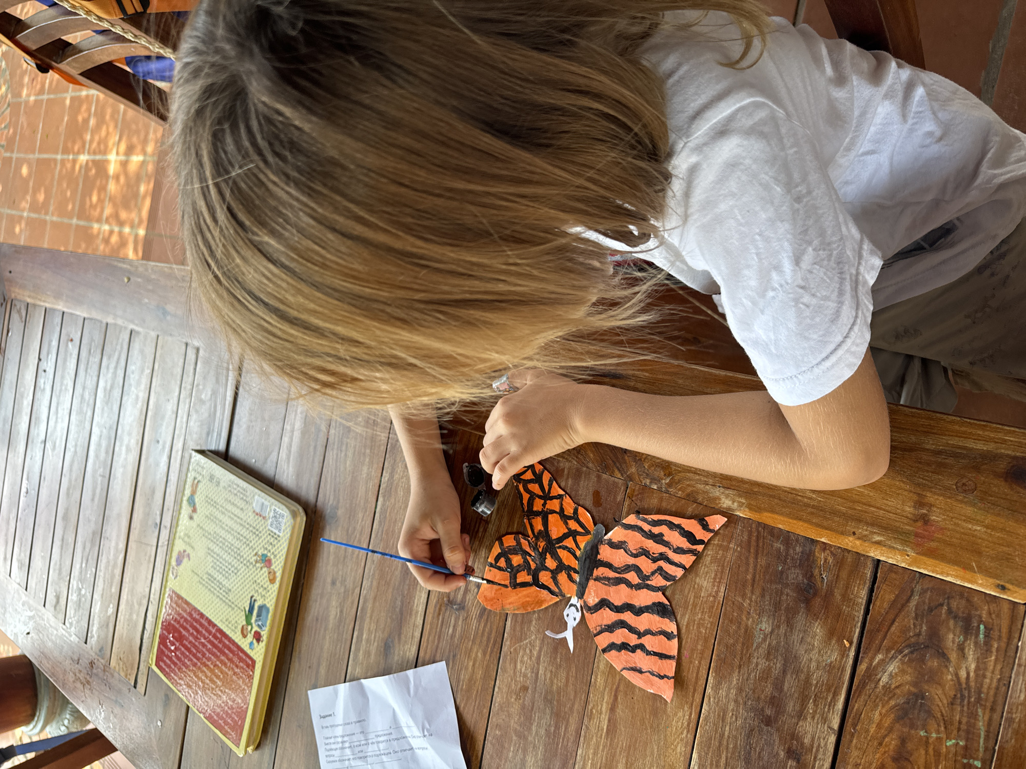 A young person, seen from an overhead perspective, is engaged in painting a craft on a slatted wooden table. The individual has long, light brown hair and is wearing a white t-shirt and light-colored pants. Their hands are focused on painting a butterfly-shaped cutout, which features an orange base with distinct black stripes, reminiscent of a tiger or monarch butterfly pattern. They hold a blue-handled paintbrush in their right hand, applying black paint from a small black pot.
On the table, to the left of the painter, lies a closed book with a yellow cover and a red spine. The back cover of the book displays illustrations and text in Cyrillic script, along with a barcode. Below the book, a sheet of white paper is visible, also containing text in Cyrillic, with the heading "Задание 1." (Task 1).
The setting appears to be outdoors or on a covered patio, suggested by the patterned shadows cast by sunlight onto a terracotta tiled floor visible in the background. The bright lighting indicates a daytime scene, likely in sunny weather. The overall scene conveys a focused, creative activity taking place in a relaxed, warm climate environment, consistent with Mũi Né, Vietnam.