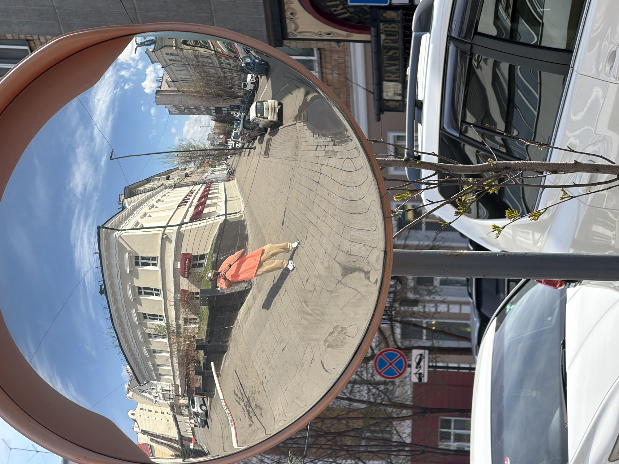 A street scene is reflected in a round convex mirror mounted on a gray metal pole, with a light brown rim. The reflection shows a city street in Krasnoyarsk, Russia. Buildings with white and beige facades and red accents line the street, and parked cars are visible in the distance. A person wearing an orange top and beige pants is visible in the reflection, standing on the brick-paved sidewalk. A "No Parking" sign is visible on the right side of the mirror's reflection. Adjacent to the mirror, parts of white cars are visible, along with a small bare tree branch with tiny leaves. A sign reading "Rat's Pub" in English is positioned above the white car. The sky is blue with scattered clouds. The overall setting is daytime and likely outdoors.