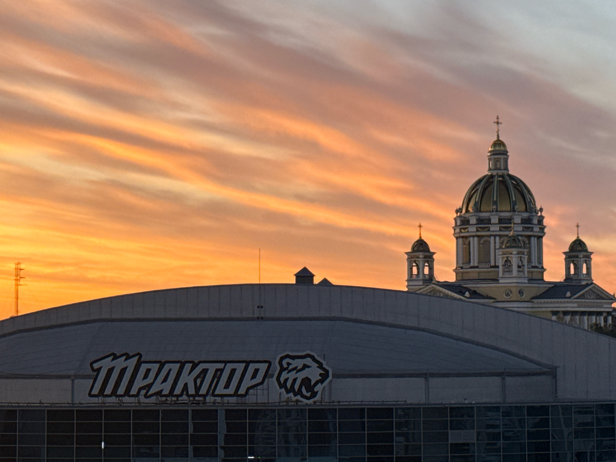The image shows a sunset over Chelyabinsk, Russia. In the foreground, a large, modern arena with the logo "TRAKTOR" and a stylized bear's head is visible. The arena has a large, curved roof and a glass facade. Behind the arena, a domed building with multiple smaller domes and crosses, resembling a cathedral or government building, stands against the colorful sky. The sky is filled with streaks of orange, pink, and yellow clouds, indicating the sun is setting. A radio tower is also visible in the distance to the left. The overall scene depicts an urban landscape with a significant architectural structure and a dramatic sky.
