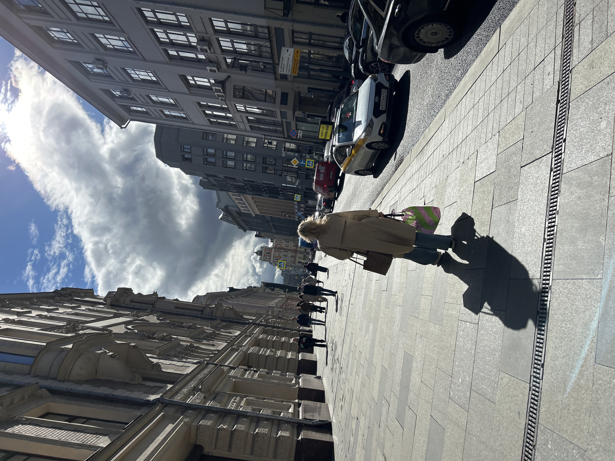 The image depicts a city street scene on a sunny day, with buildings lining both sides of the street. Several people are walking along the sidewalk, and cars are parked along the curb. A woman wearing a beige coat and carrying a pink and green bag walks prominently along the sidewalk, with her shadow stretching beside her. The pavement is made of rectangular stones, and there is a metal grate running along the edge. The sky is partly cloudy, with patches of blue visible. Street signs are visible attached to buildings. The location is Unknown, Unknown.