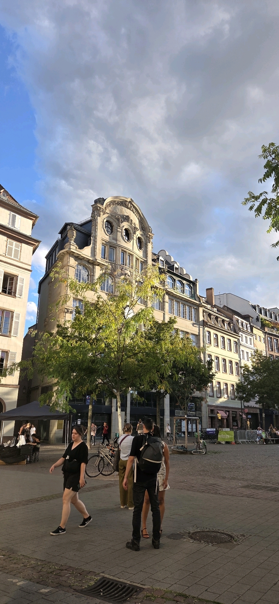 The image depicts a street scene in Strasbourg, France, likely in a historic district given the architecture. In the foreground, a woman in a black t-shirt and shorts walks from left to right, while a man with a backpack walks away from the camera. Behind them, several other people are visible, some walking and others standing. A tree with green foliage is prominently featured in the mid-ground, casting dappled sunlight on the pavement.
The background is dominated by a multi-story building with ornate architectural details, including arched windows and decorative elements on the facade. The building appears to be a mix of residential and commercial spaces, with shopfronts visible on the ground floor. Adjacent buildings line the street, showcasing similar historical architecture with multiple windows and pitched roofs. The sky above is partly cloudy with patches of blue sky and darker gray clouds, suggesting a dynamic weather pattern. The ground is paved with cobblestones and a metal grate is visible in the lower foreground. Bicycles are parked in various locations, indicating a mode of transportation used in the area. The overall atmosphere suggests a lively public square or street.
