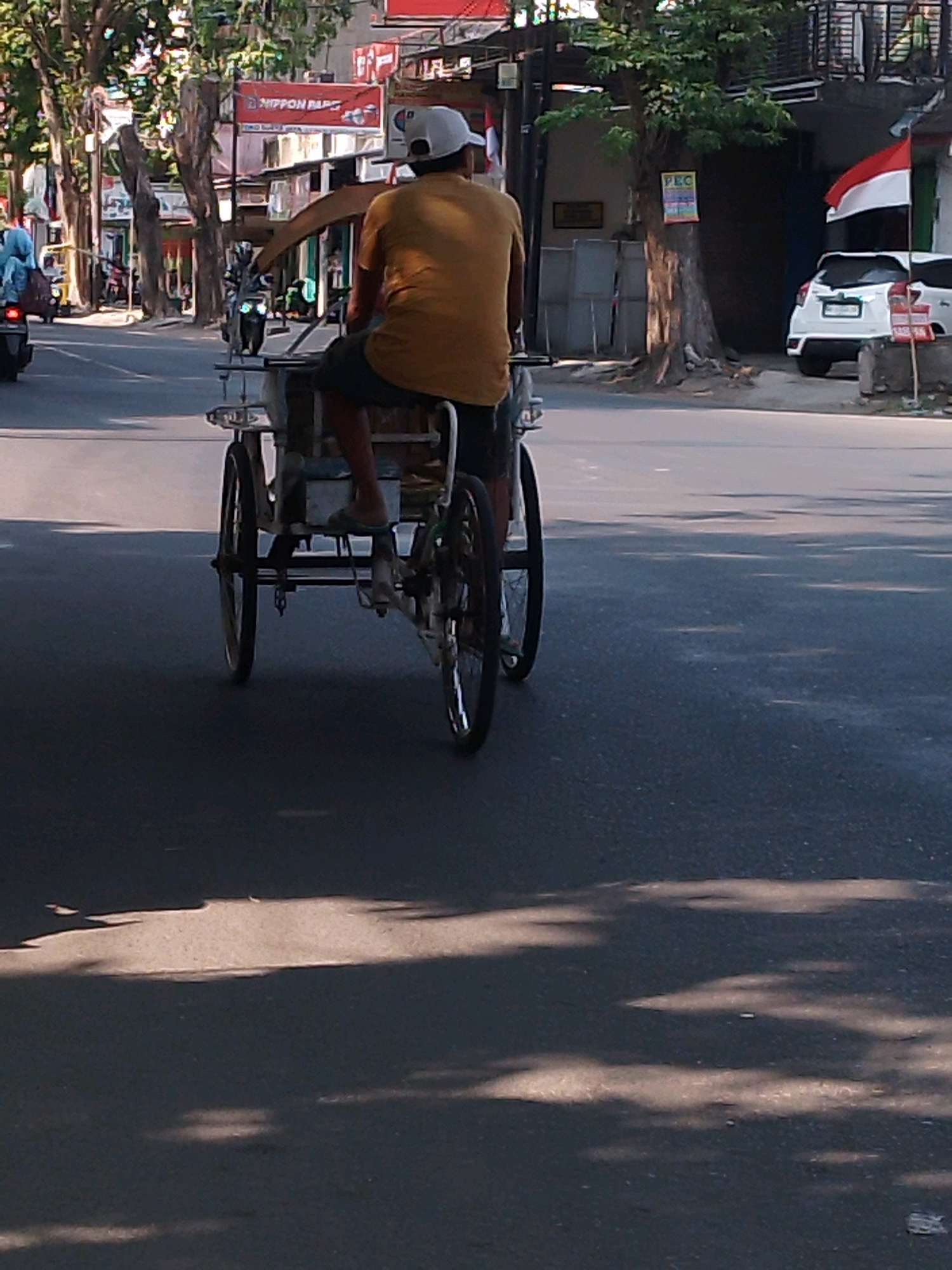 The image shows a becak driver, a three-wheeled passenger vehicle common in Indonesia, on a street in Kertosono. The driver, wearing a yellow t-shirt and a white cap, is seated in the rear of the becak, steering it. The becak has a canopy and seating for passengers. In the background, there are buildings, trees, and other vehicles, including a white car parked on the side of the road. The scene appears to be in an urban or semi-urban area, likely during daylight hours. A red and white flag is visible on a pole near the parked car.