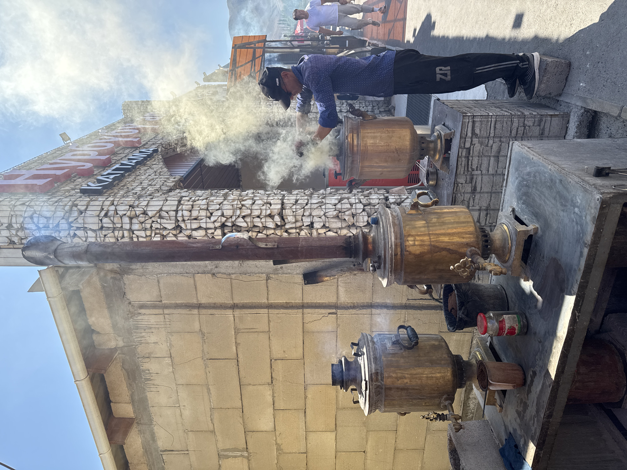 In Sasyk-Bulak, Kyrgyzstan, on a sunny day, a man wearing a blue patterned shirt, black cap, and black pants with a "ZR" logo is tending to a steaming samovar. Two large, ornate brass samovars are visible, with the man actively working at the larger one, which is emitting a significant amount of steam. Next to the samovars are various items including a metal pitcher, a jar of what appears to be jam, and a black trash bin. The scene takes place in an outdoor area with a wall constructed of light-colored bricks. Behind the man, a building with a distinctive facade of stacked stones and a large red sign with white lettering is visible, along with a smaller black sign that reads "KATTANYA." Another man in a white t-shirt and light-colored pants walks in the background, suggesting a public or commercial setting. The overall environment appears to be a street or an open-air establishment.