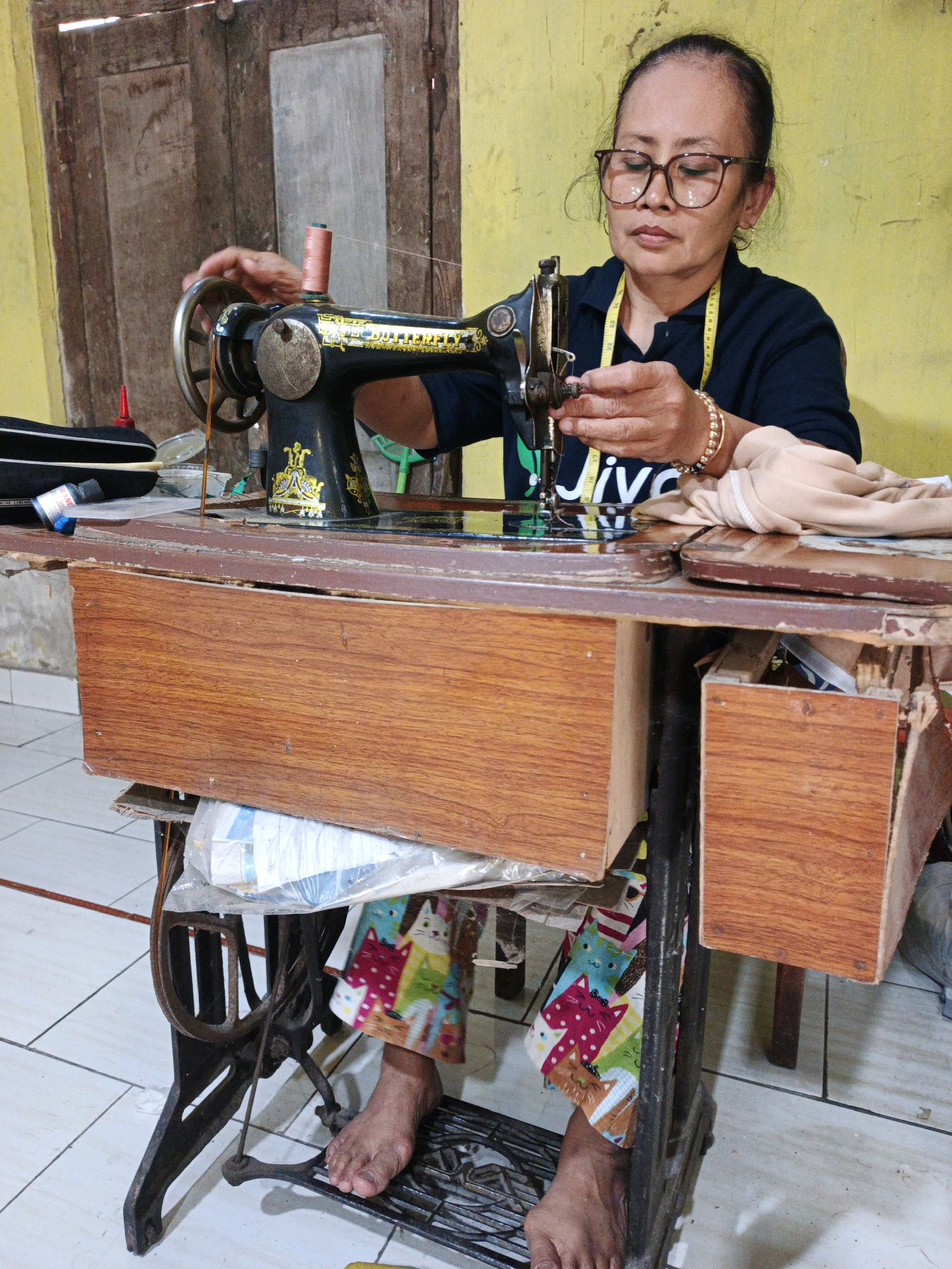 A woman is operating a vintage sewing machine in Modongan, Indonesia. The sewing machine is a black, ornate model with gold detailing, positioned on a wooden table with drawers. The woman, wearing glasses and a dark blue shirt, has a yellow measuring tape around her neck. She is actively sewing, with her hands engaged with the needle and fabric. The surrounding environment appears to be an indoor space with a yellow wall and a wooden door in the background. The floor is tiled. The woman's lower legs are visible, and she is wearing colorful pants with a cat print. Her bare feet are positioned on the sewing machine's treadle.