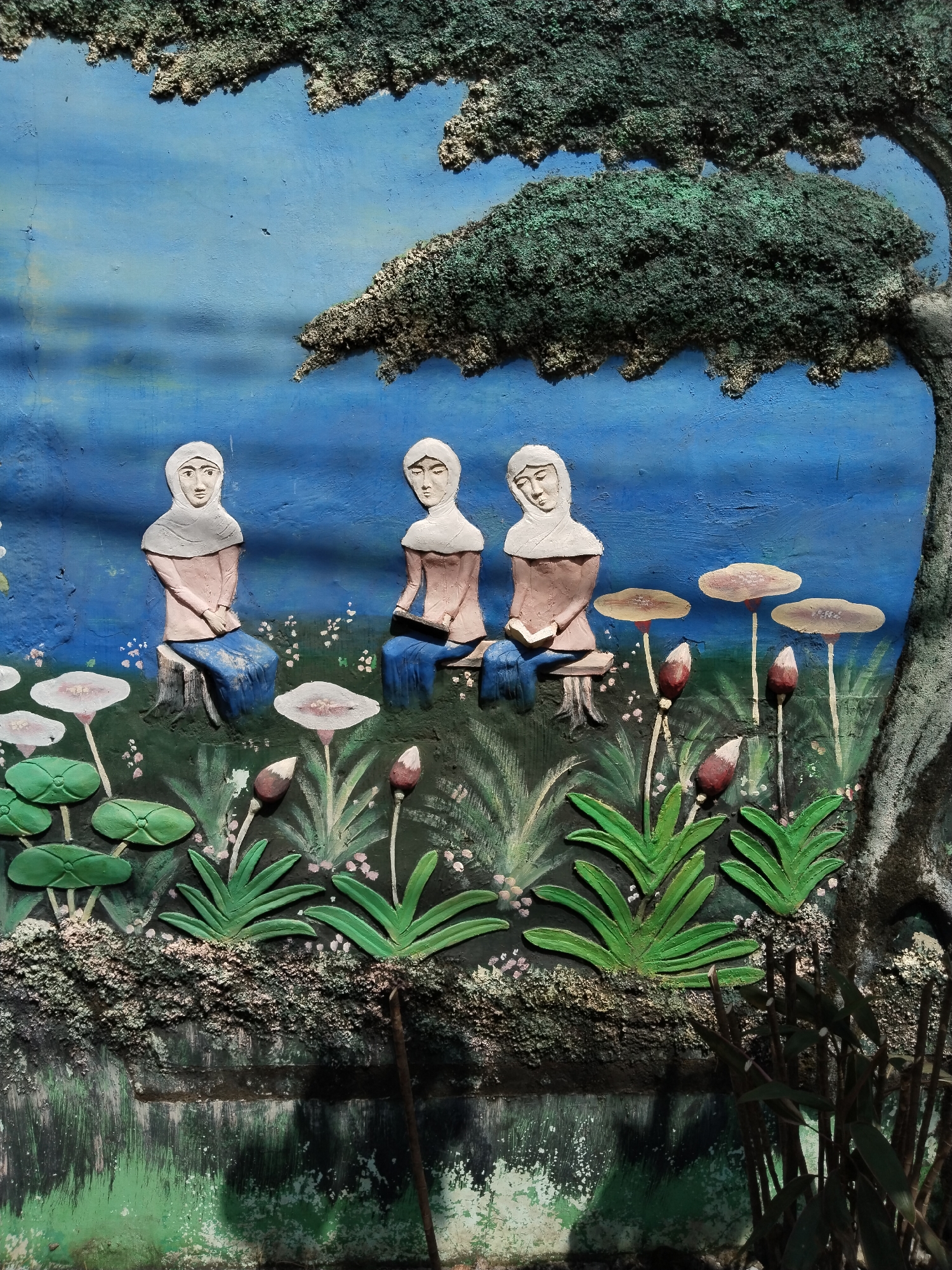 This media file captures a detailed, colorful bas-relief mural, likely depicting a serene outdoor scene in Sepat, Indonesia.
The primary subjects are three female figures, all wearing white hijabs and modest clothing (light brown tops, blue skirts/drapery). They are seated in a line, carved in relief, with the figure on the far left sitting on a tree stump, and the other two appearing to be on simple benches. The figure on the far right is holding what appears to be a book or tablet, suggesting an activity of reading or studying. Their expressions are calm and contemplative.
The setting is a lush natural environment. A large tree with thick, textured dark green foliage frames the scene on the upper right, its trunk extending down the right edge. The background is a vibrant blue, possibly representing a sky or a distant body of water. The foreground features various carved plants and flowers, including broad green leaves resembling water lilies or lotus pads, several light-colored mushroom-like structures, and reddish-brown flower buds on stems. The ground itself is highly textured, with rough, mossy-looking patches and painted details of smaller plants. Below this, a body of water is depicted, with reflections and painted light patches, suggesting a pond or river at the base of the scene.
The overall artwork is three-dimensional, with painted details enhancing the sculpted forms. The textures are particularly noticeable on the tree's foliage and the ground, giving the mural a tactile quality. The activity portrayed suggests a peaceful gathering, possibly for study or reflection amidst nature. There is no visible text in the image.