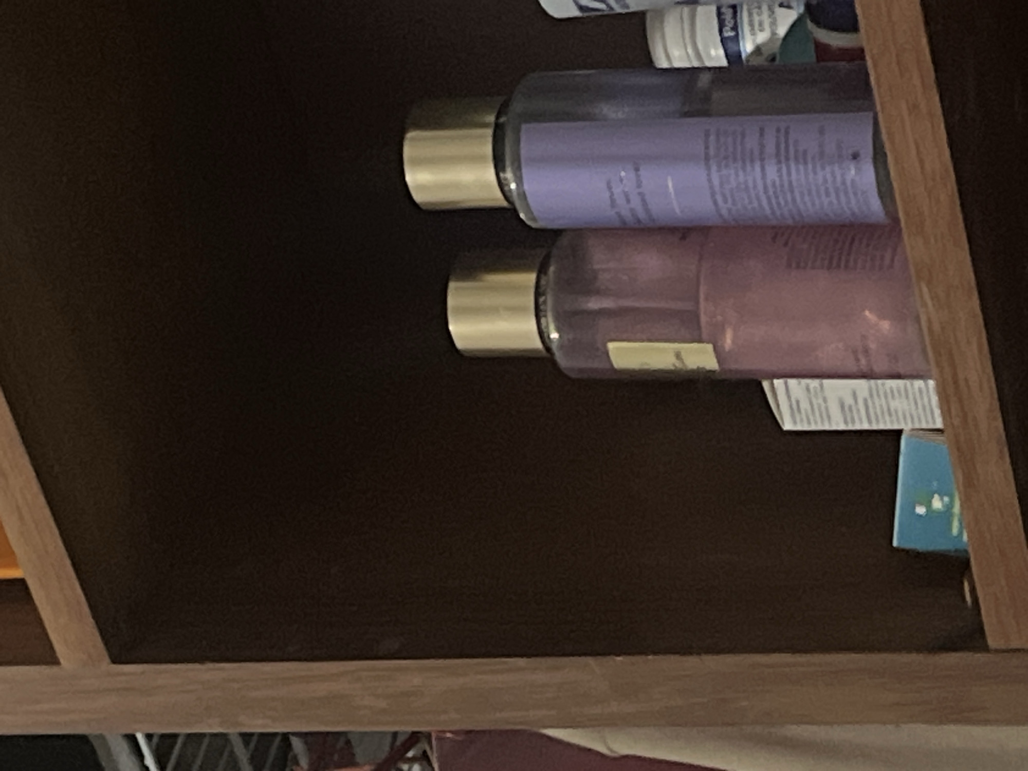 The image shows a close-up view of two clear plastic bottles with gold caps sitting on a wooden shelf. The bottle on the left contains a pink liquid, and the bottle on the right contains a purple liquid with a label that has text on it. To the right of these bottles, a white and blue bottle is partially visible, with the word "Peld" on its label. In the foreground, a small blue box with white markings is positioned on the shelf. The background is dark and out of focus, suggesting a dimly lit environment. There are no people or any indication of the city of Santa Ana, Venezuela present in the image.