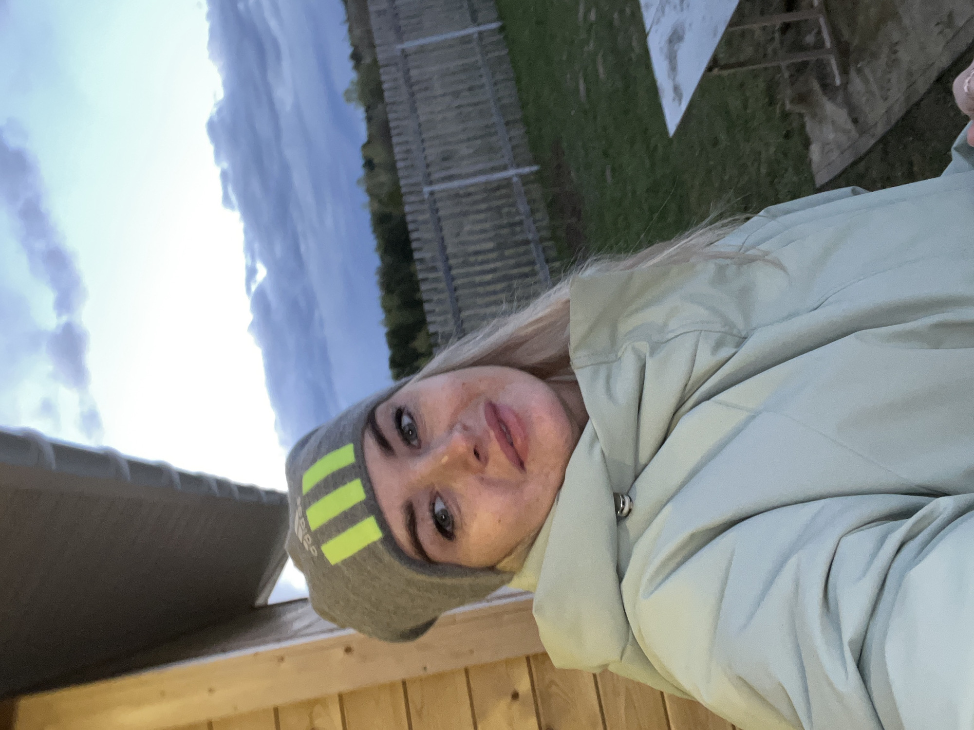 A woman is posing for a selfie outdoors. She is wearing a light green puffer jacket and a gray beanie with yellow stripes. The sky behind her is cloudy, transitioning from light to dark. Behind her, there's a wooden fence and green grass. A building's wooden roof and wall are visible in the foreground. The location is Гаревая, Russia.