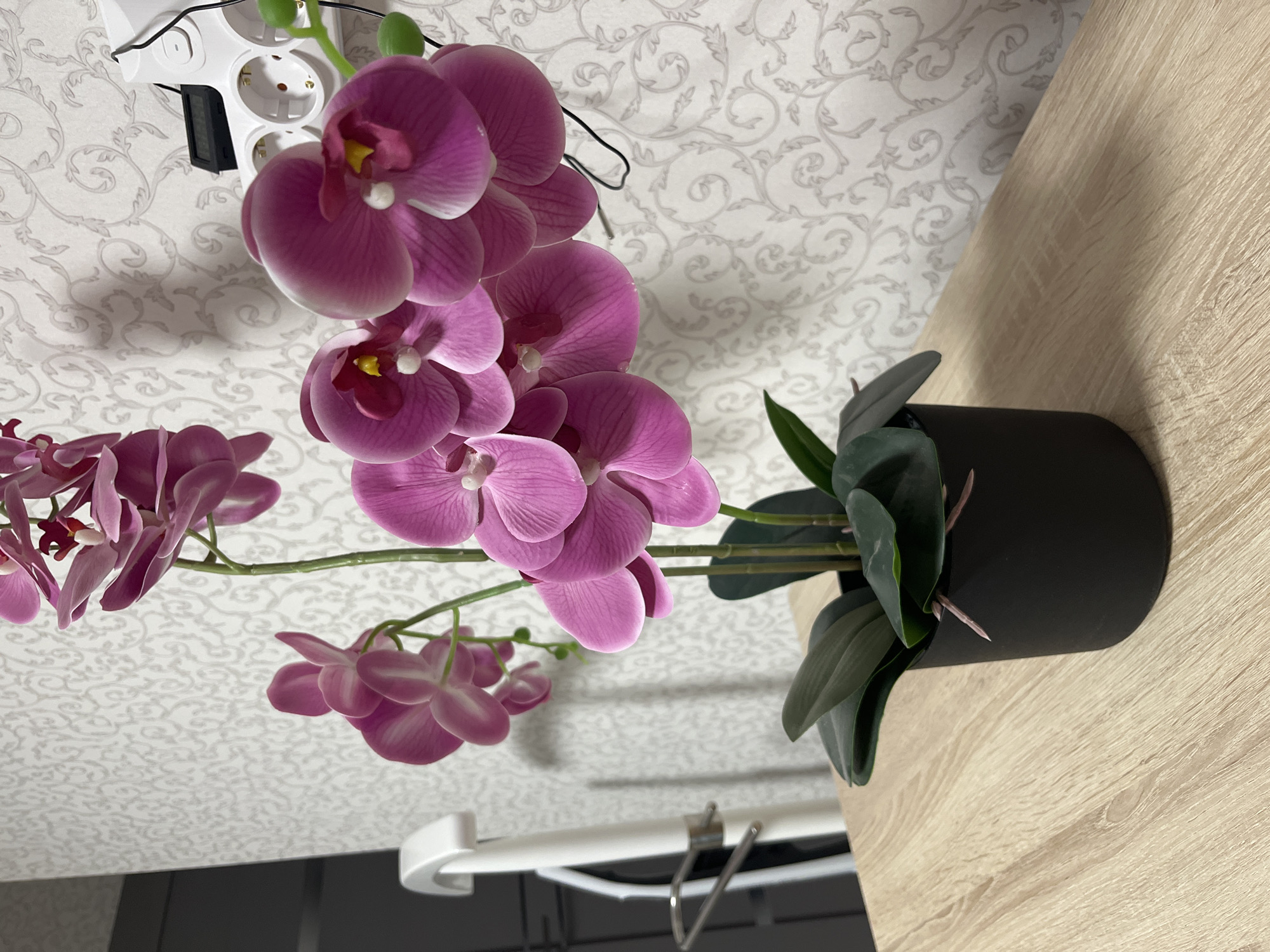 This image presents a close-up view of an artificial orchid plant situated on a light-colored wood-grain surface, likely a table or countertop. The plant is contained within a matte black cylindrical pot and features multiple fuchsia-pink faux orchid blossoms arranged on green stems, accompanied by several dark green artificial leaves at the base. The background comprises a light-colored wall adorned with a subtly patterned wallpaper featuring a repeating, ornate, light grey scroll or floral design. To the upper right, a white multi-socket power strip, containing three visible outlets, is mounted on the wall. A small black digital device, possibly a thermometer or hygrometer, is positioned on the top of the power strip, with a black cable extending from it. A portion of a white, curved furniture element is partially visible on the far left of the image. The scene is located in the city of Старые Кены, Russia.
