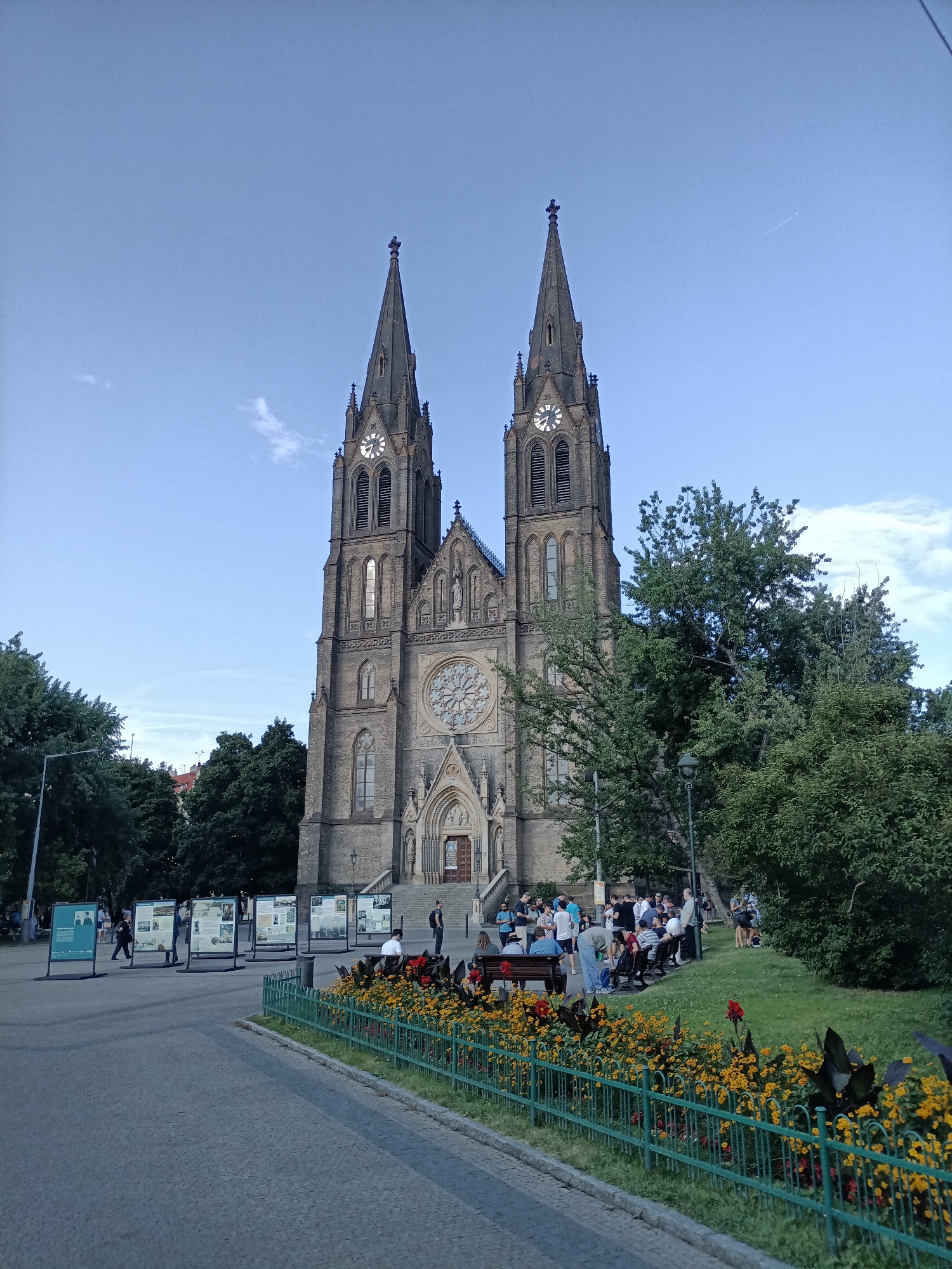 This image depicts the Basilica of Saint Peter and Saint Paul in Vyšehrad, Prague, Czechia. The large gothic cathedral is the central subject, with its twin spires prominently reaching towards a clear blue sky. Several people are gathered in the foreground on a grassy area and on benches, appearing to be resting or observing the architecture. Others are standing near information displays set up on the plaza in front of the church. The environment includes lush green trees flanking the church and surrounding the gathering area. A vibrant flowerbed with yellow and red flowers lines a green metal fence in the foreground, adding a touch of color to the scene. The overall atmosphere suggests a public space in a city, with a blend of historical architecture, recreational activity, and natural elements.