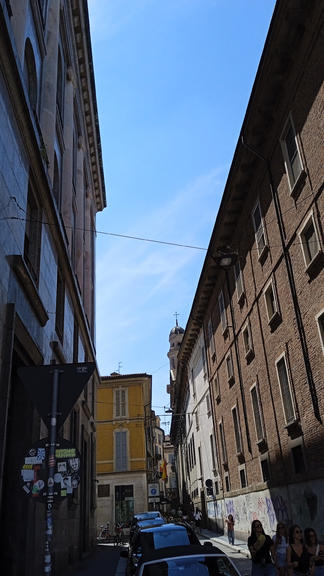 The image captures a street scene in Milan, Italy, likely in an older district. The perspective is looking up a narrow street flanked by buildings on both sides. On the left, a building with arched windows and a stone facade dominates the foreground, casting a shadow. A traffic sign with various stickers is mounted on a pole in front of this building.
On the right, a taller brick building with numerous windows runs along the street. The building's facade shows signs of age, with some graffiti visible at its base. Further down the street, a yellow building with shuttered windows is visible, and beyond that, more buildings, some appearing older and with distinct architectural features like a domed tower topped with a cross, suggesting a church or campanile.
Several cars are parked along the street, including a black convertible in the foreground and other vehicles further down. A few people are scattered along the sidewalk, some walking and others standing in groups. A group of women are visible near the right side of the image, some of them looking towards the camera. The sky is clear and blue with a few wispy clouds. The lighting suggests it is daytime, with strong shadows indicating bright sunlight. The overall impression is of a European city street with a mix of old and new elements, bustling with daily activity.