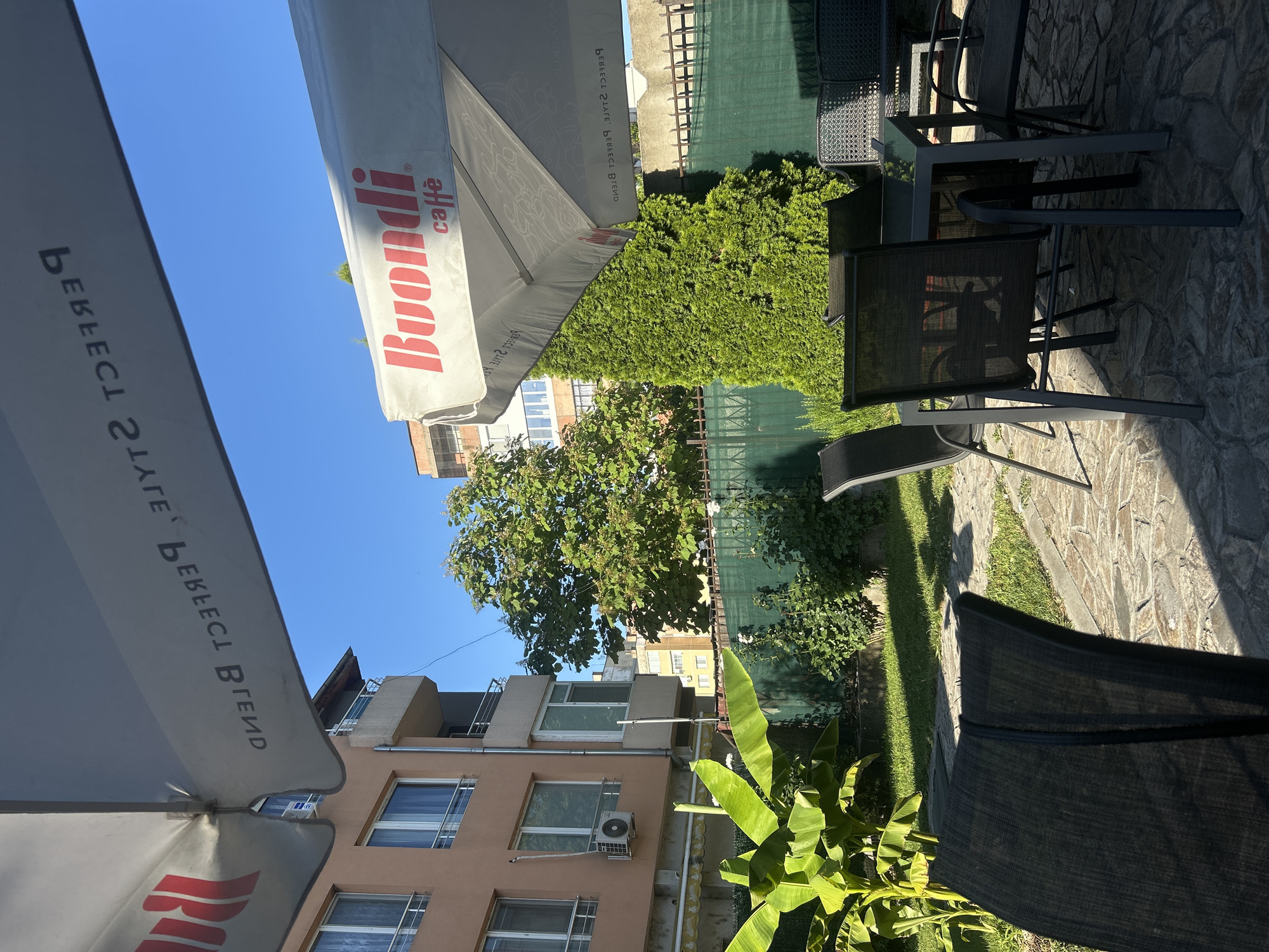 The image depicts an outdoor seating area at a cafe, likely located in Vidin, Bulgaria. In the foreground, several dark-colored chairs and a table are arranged on a stone-tiled patio. A large umbrella, adorned with the "Buondi Caffè" logo, is partially visible, casting a shadow over the seating area. In the background, a building with a light brown facade and balconies stands adjacent to lush green foliage and a tall, conical evergreen tree. The scene is bathed in sunlight, indicating it is daytime. The overall impression is that of a tranquil outdoor dining space.