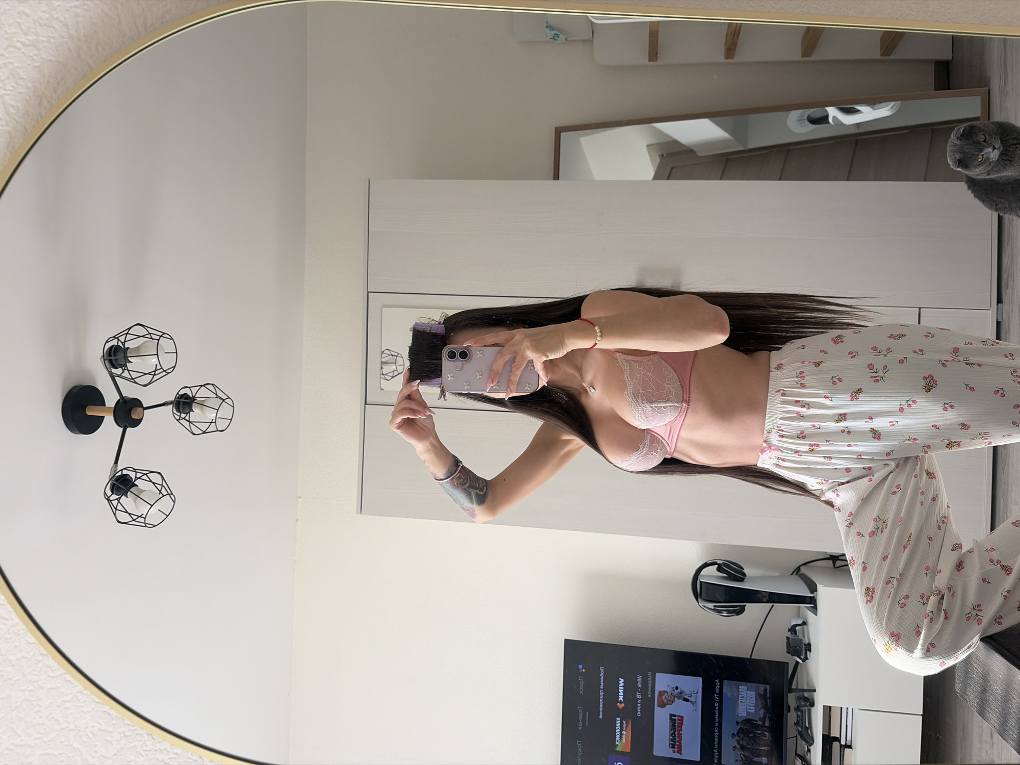 A mirror selfie of a woman indoors. She wears a pink bra and floral pajamas. She holds a phone, presumably taking the picture. A cat sits in the background, and there's a television and gaming console nearby. The background suggests a modern interior.