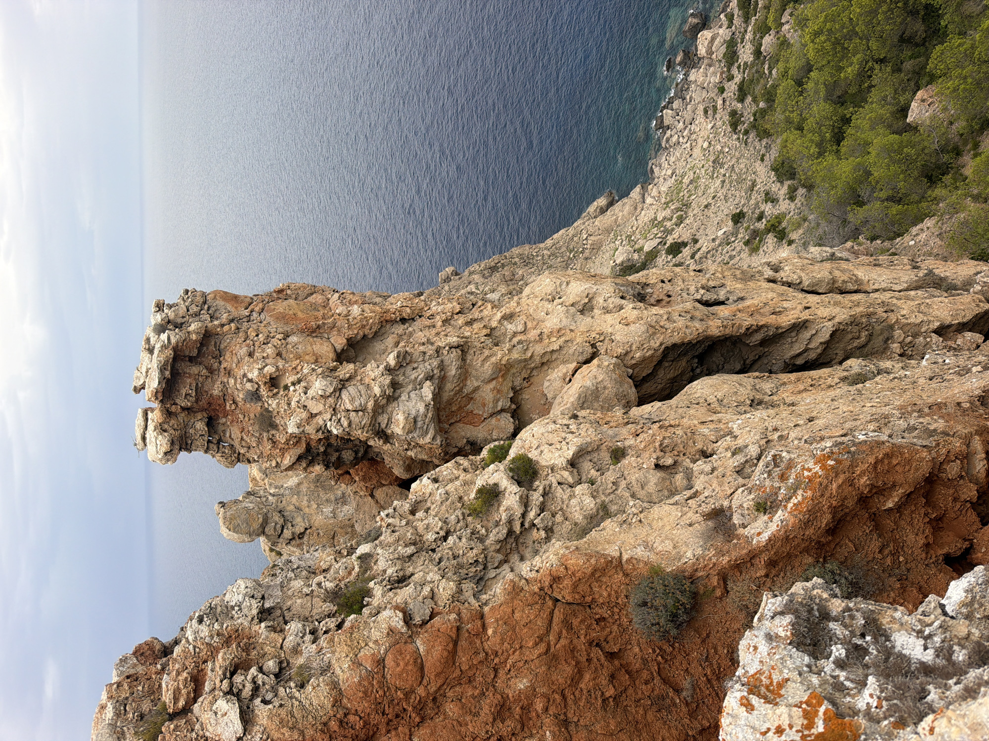 The image shows a view from a rocky cliff overlooking the sea near Sant Antoni de Portmany, Spain. The cliff face is composed of layered rock formations with varying textures and colors, ranging from reddish-brown to light tan. Patches of sparse, green vegetation are visible clinging to the rocks. The sea is a deep blue, with small waves creating a textured surface. The sky is overcast with a pale gray hue. The scene is natural and undeveloped, with no visible human presence or structures.
