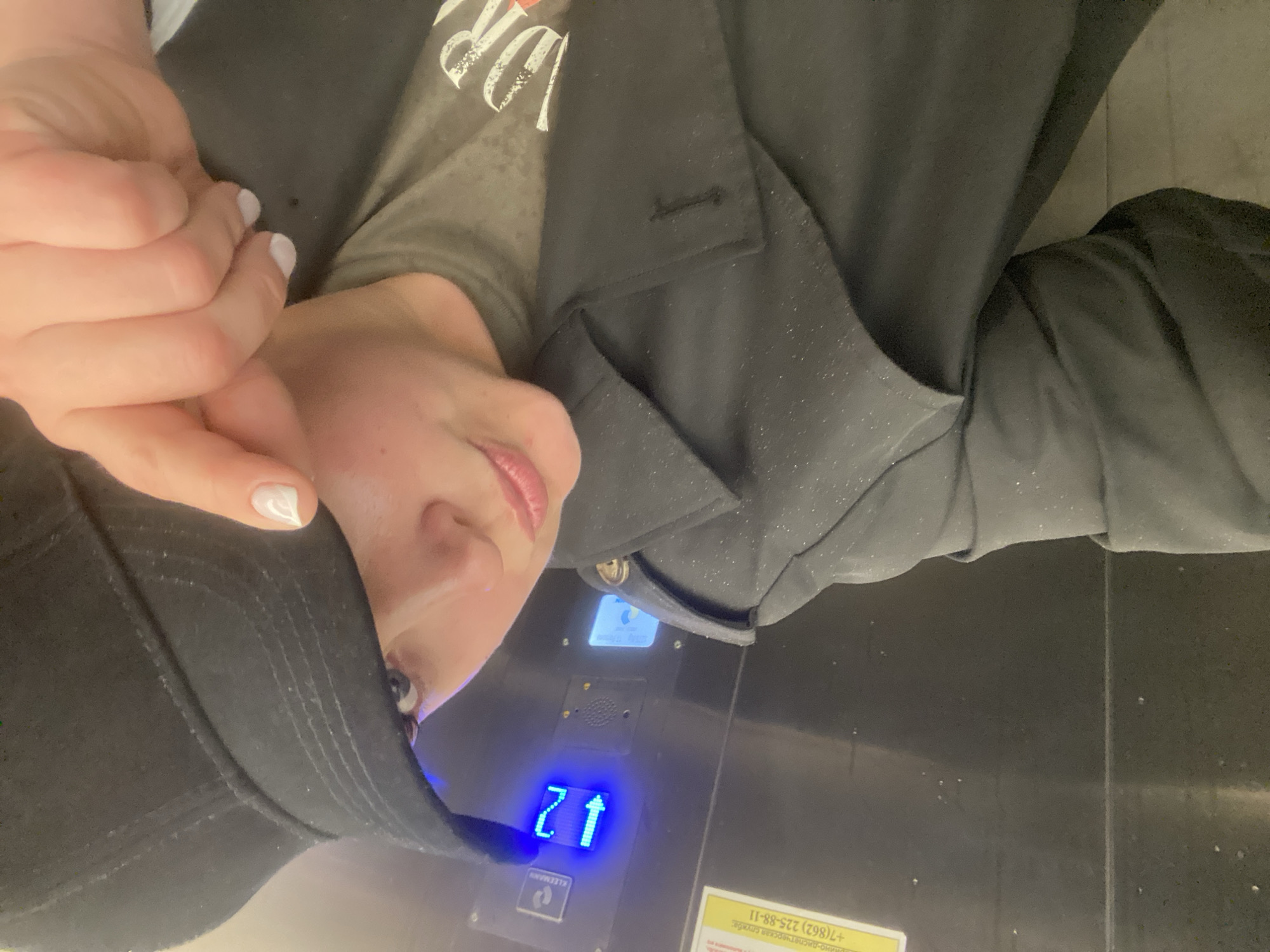 A woman wearing a black baseball cap, grey t-shirt, and black blazer is posing for a photo in an elevator. The elevator's floor indicator displays "21" with an upward arrow, suggesting it is moving upwards. The elevator is in Sochi, Russia, as indicated by a sign with the phone number +7(862) 225-88-11.