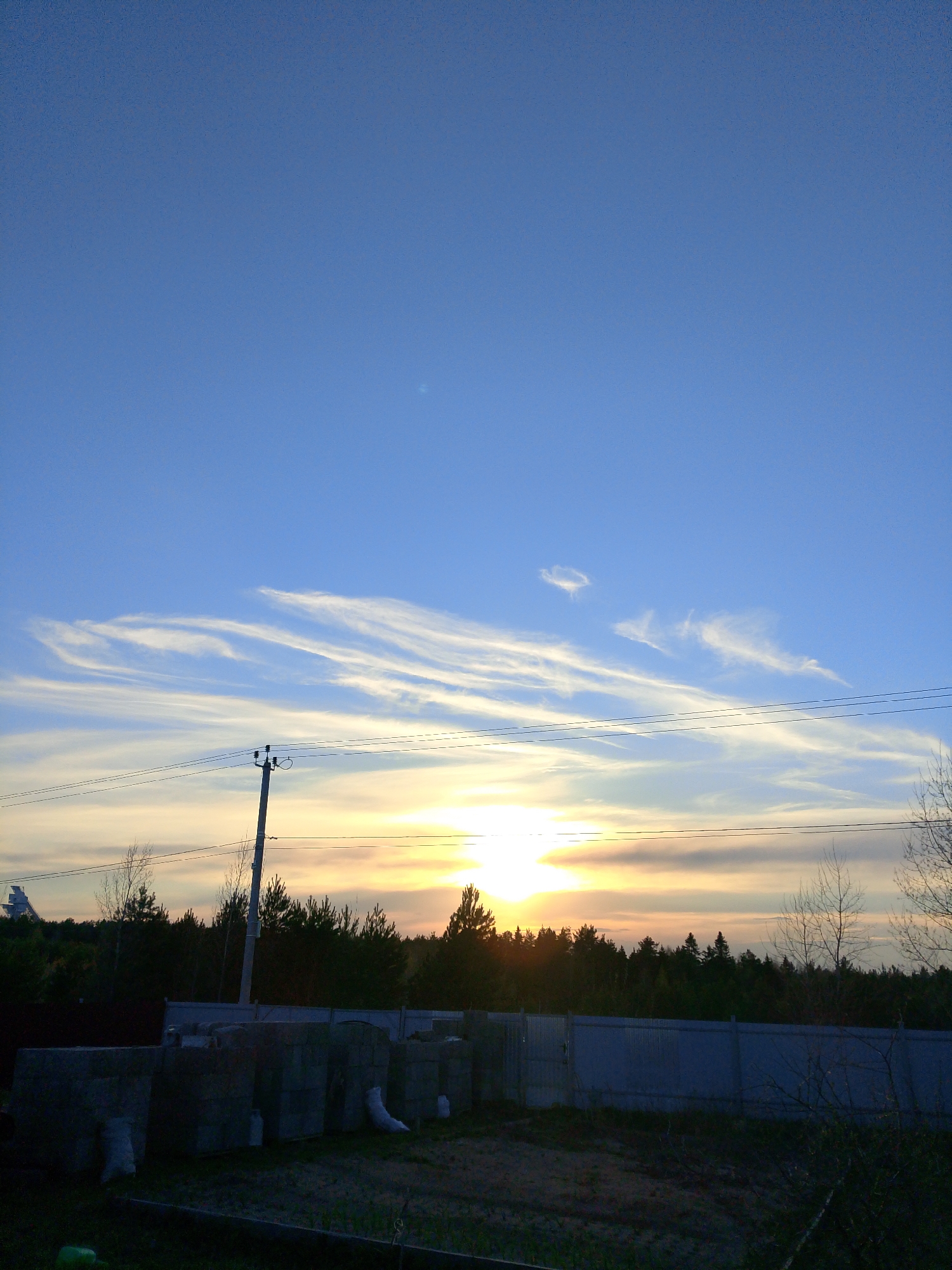 The image depicts a scene in Прикамский, Russia, featuring a landscape at dusk or dawn. In the foreground, there is a plot of land with construction materials, including concrete blocks and bags, partially obscuring a fence. The middle ground shows a line of trees silhouetted against a brightly lit horizon, indicating the sun is either setting or rising. A utility pole with power lines extends across the scene. The sky transitions from a deep blue at the top of the frame to a pale yellow-orange near the horizon, with streaks of white clouds.