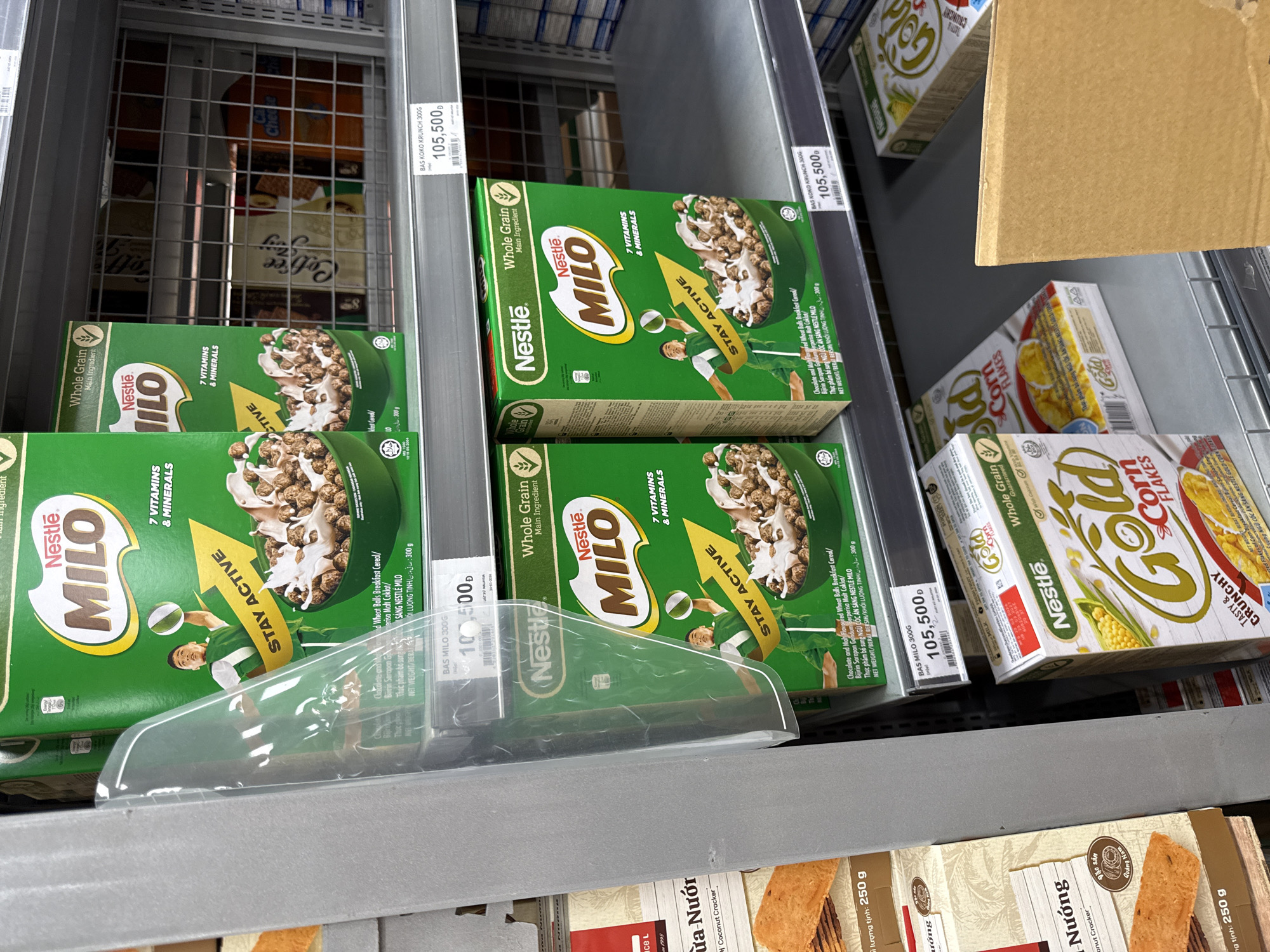 A close-up shot on a supermarket shelf in Nha Trang, Vietnam, displays rows of breakfast cereal boxes. The dominant items are green boxes of Nestle Milo, with the "STAY ACTIVE" slogan and a depiction of a young man playing a sport. To the left of the Milo boxes, partially visible, are boxes of "An An Nuong Coconut Cracker". On the right side of the shelf, the boxes continue with more Milo and Nestle Gold Corn Flakes. The lower shelf shows a similar arrangement of cereals, including Gold Corn Flakes. The lighting suggests an indoor retail environment.