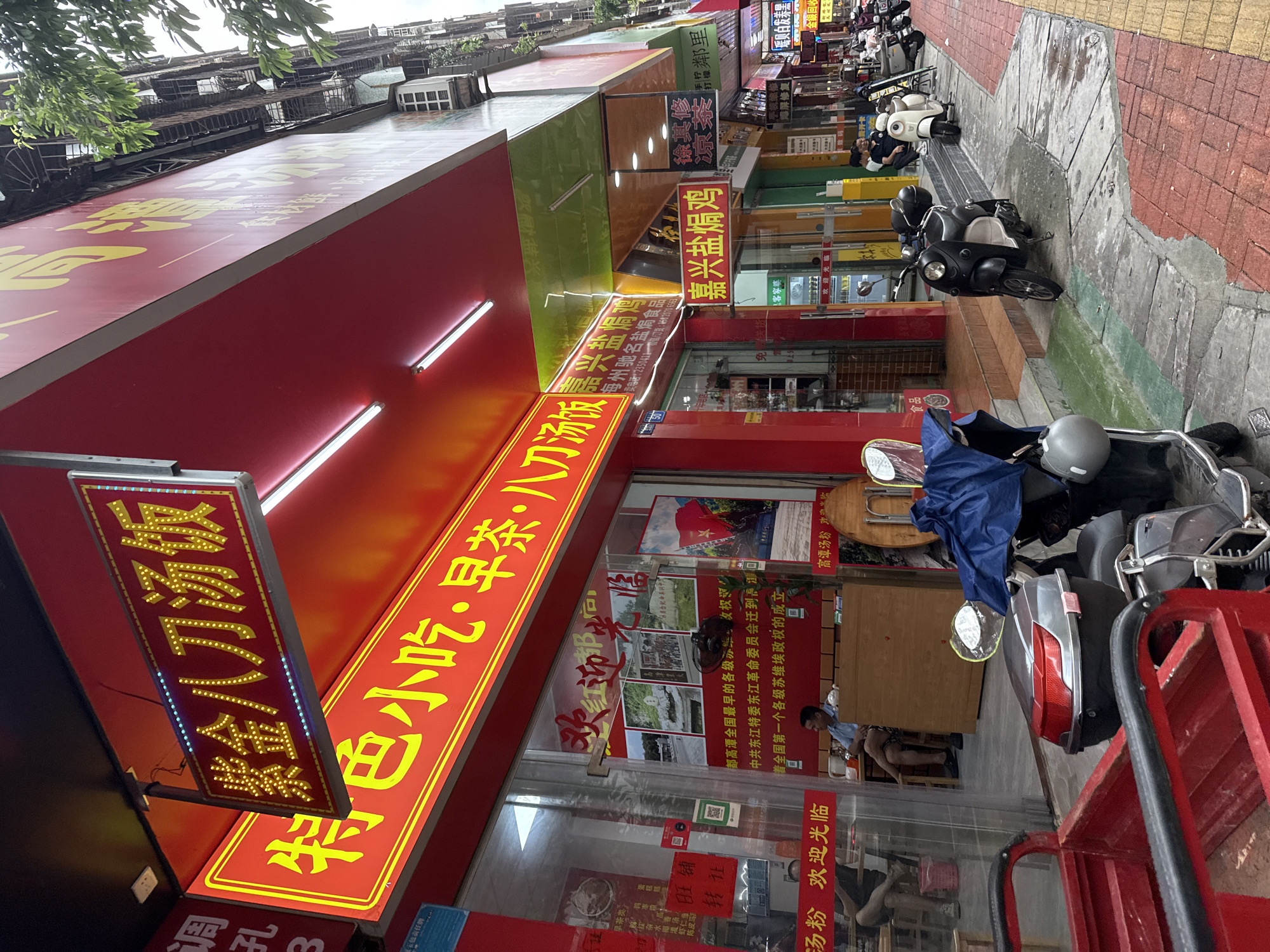 A street scene in Huizhou, China, captures the bustling atmosphere of a commercial district with a focus on food establishments. The foreground is dominated by a red three-wheeled vehicle, possibly a cargo tricycle, with its front end partially visible. To its right, a dark-colored scooter is parked, covered partially by a blue tarp, with a helmet attached to its rear. Behind these vehicles, the facade of several businesses lines the street.
Prominently displayed on the left is a red awning with prominent yellow Chinese characters indicating a food establishment. A sign above reads "紫金八刀汤饭" (Zijin Ba Dao Tang Fan), suggesting a specialty dish. Below this, another red sign with yellow lettering says "特色小吃·早茶·八刀汤饭" (Specialty Snacks, Morning Tea, Ba Dao Tang Fan), further detailing the offerings. The shopfront features large glass windows, through which customers can be seen sitting at tables, enjoying meals.
Further down the street, other businesses are visible, each with their own signage. One sign, illuminated in yellow against a red background, reads "嘉兴盐焗鸡" (Jiaxing Salt Baked Chicken). Another, with green lettering on a white background, has Chinese characters that are partially obscured but suggest it is also a food or beverage establishment. Various motorcycles and scooters are parked along the sidewalk, indicating common modes of transportation in the area. The buildings on the right are multi-story structures, typical of urban environments, with visible air conditioning units and balconies. The sidewalk is paved with a mix of red bricks and grey concrete, with yellow lines marking some sections. The overall impression is one of a lively street, active with commerce and daily life in Huizhou.