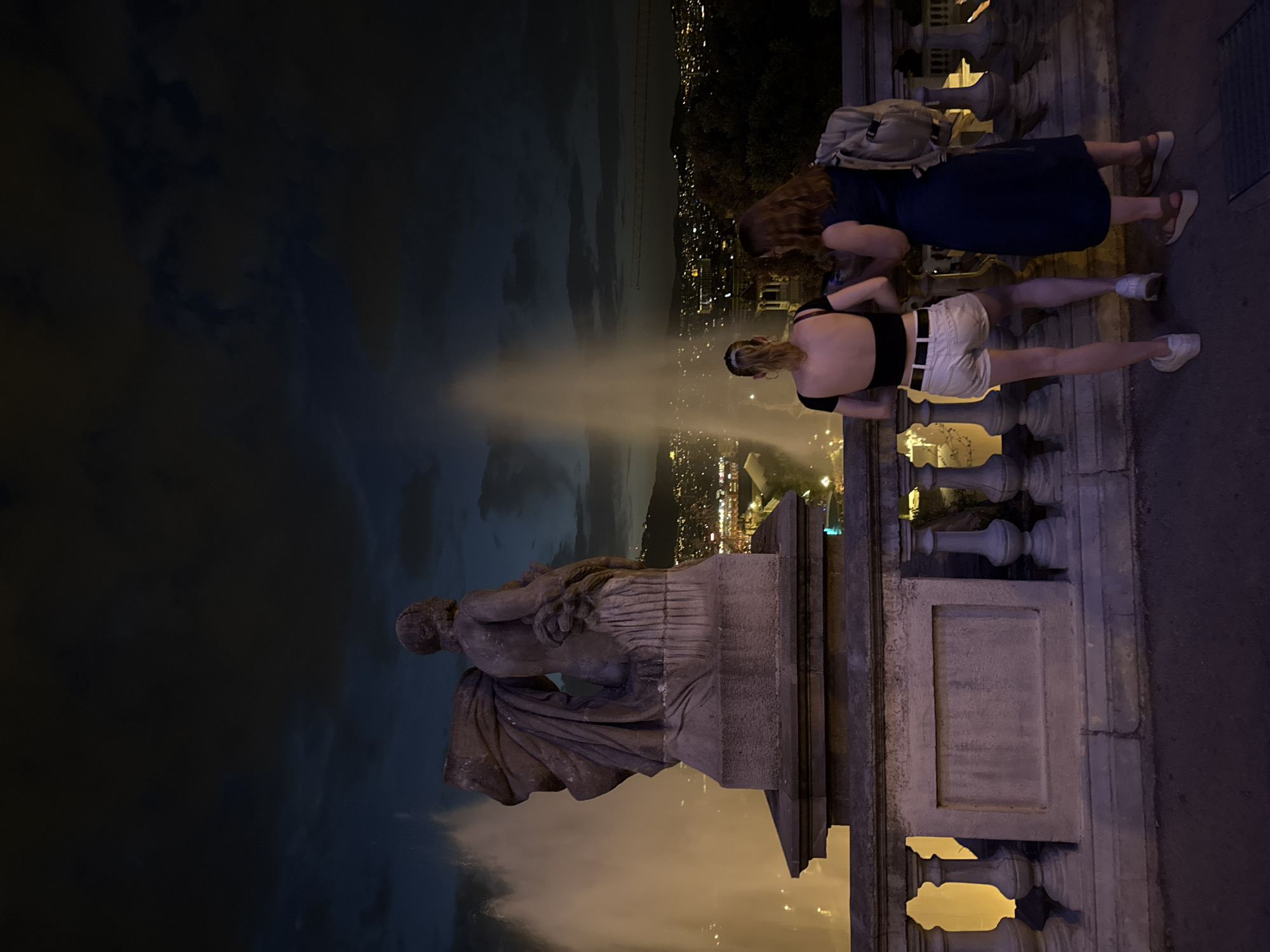 The image shows two women standing on a stone balcony at night, overlooking a city illuminated by lights. Behind them, a large fountain is spraying water, creating tall plumes. In the foreground, a stone statue of a figure with draped clothing stands on a pedestal. The women are facing away from the camera, looking towards the city lights and the fountain. The woman on the left is wearing a black crop top and white shorts, while the woman on the right is wearing a dark blue dress and a backpack. The sky above is dark with some clouds. This scene is likely from the Magic Fountain of Montjuïc in Barcelona, Spain, given its distinctive fountain and panoramic city views, often enjoyed at night.