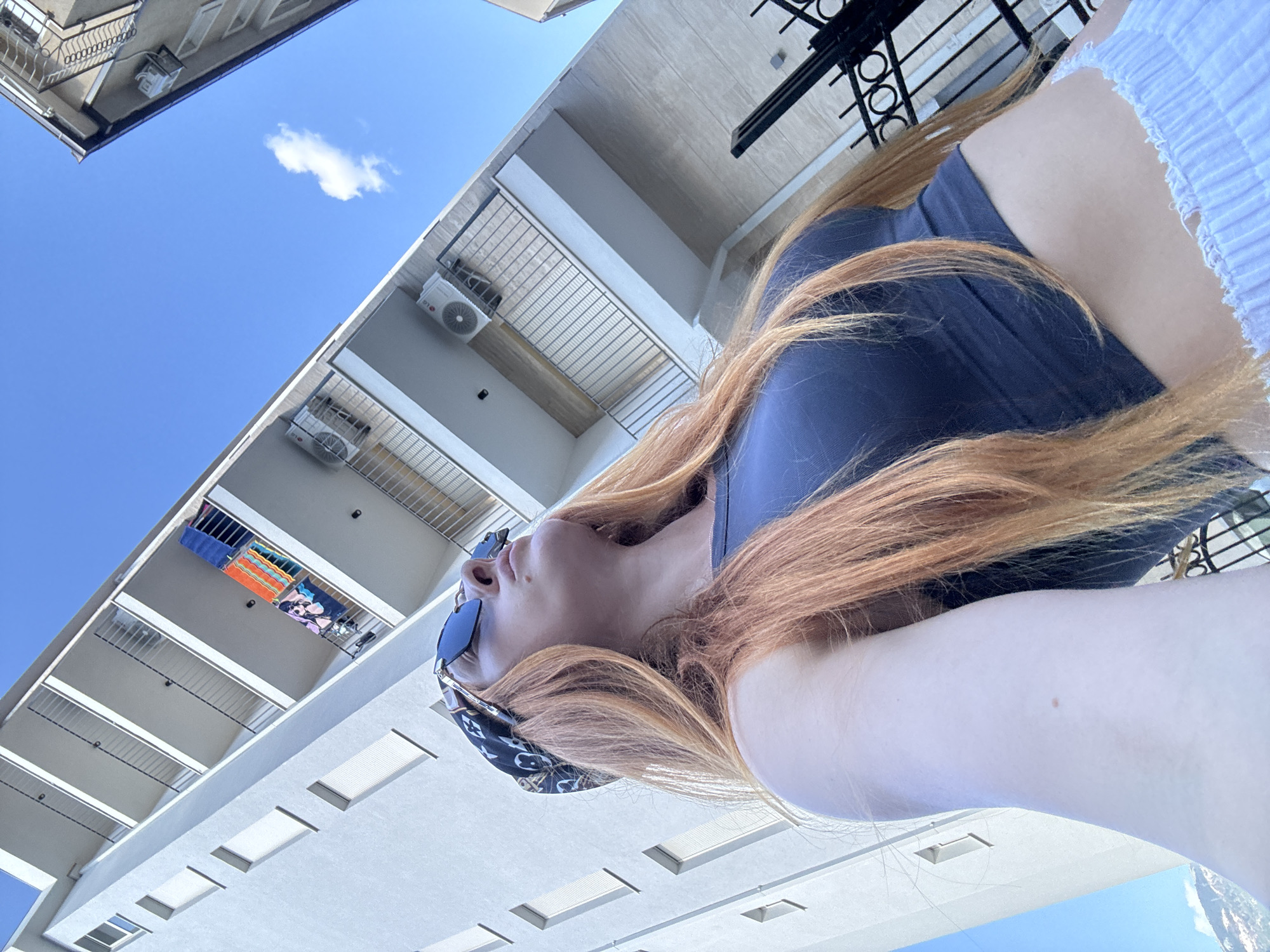 The image shows a woman with long, red hair, wearing sunglasses and a bandana. She is wearing a dark tank top and white shorts. The woman is looking up towards a building with multiple balconies. The building is white and appears to be an apartment building. There are air conditioning units on some of the balconies, as well as towels and clothing. The sky is blue with a few clouds. The image appears to have been taken in Budva, Montenegro, as indicated by the presence of the building which is located on or near the Budva Riviera.