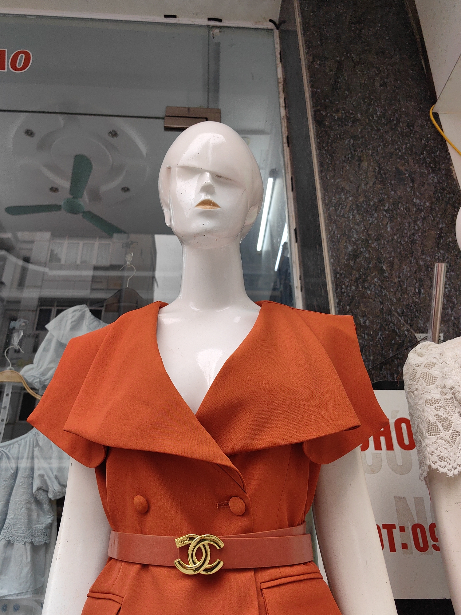 A mannequin in Bai Chay Ward, Vietnam, wears an orange blazer-style dress with a tan belt and a gold-colored double-C logo buckle. The mannequin is displayed in a storefront window, with a reflection of a ceiling fan and buildings visible on the glass. A second mannequin wearing white lace is partially visible to the right. A sign with the text "HO" and "T:09" is behind the mannequins. A gray stone-like surface forms the edge of the storefront.