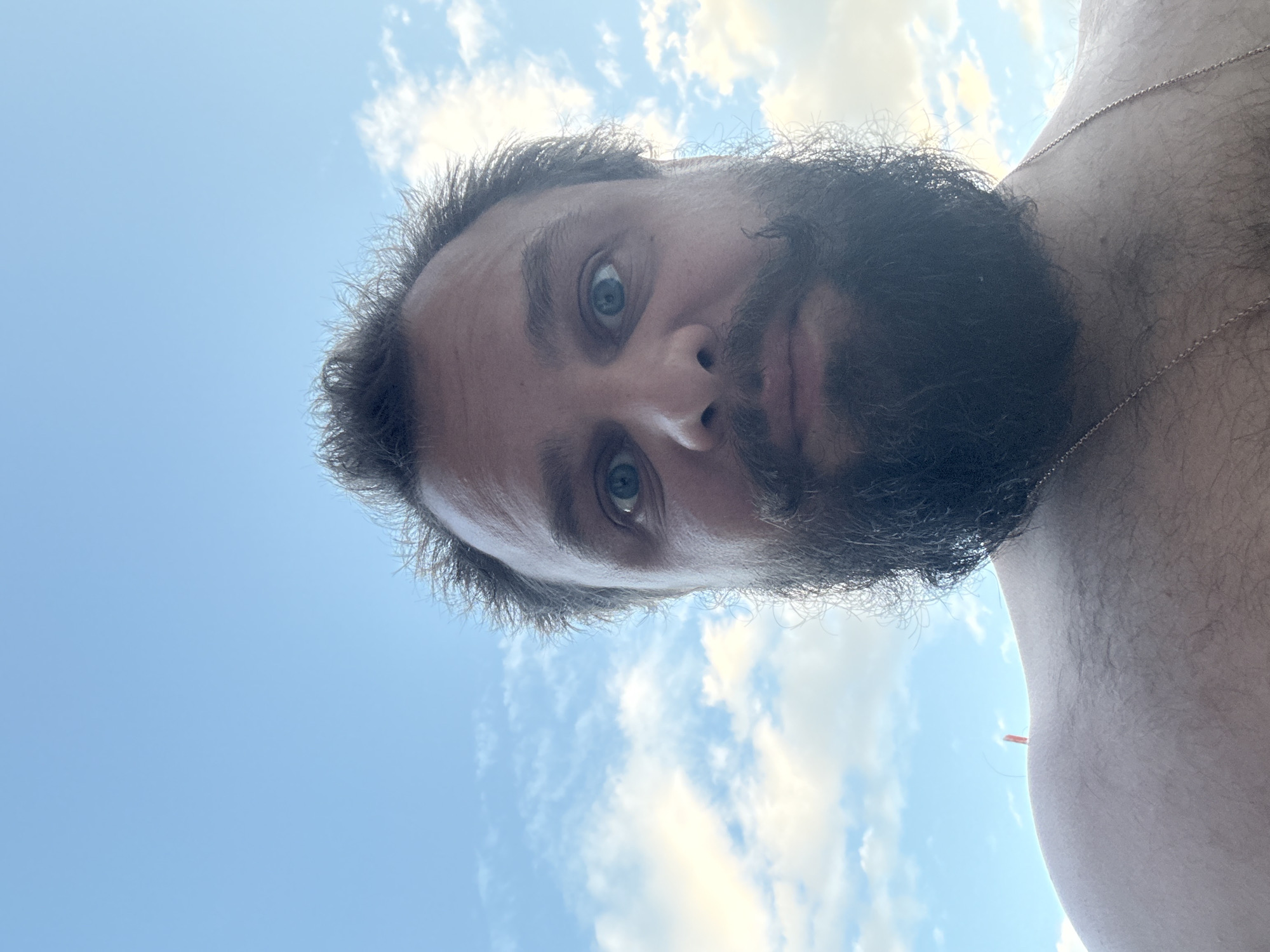 An extreme close-up selfie of a man's face and upper torso is captured against a bright blue sky with scattered white clouds. The man has dark, somewhat unkempt hair, prominent eyebrows, and wide, blue eyes. He sports a full, dark beard and mustache. The camera angle is from below, looking upwards at the man. He appears to be shirtless, with a faint glimpse of a thin gold chain around his neck. In the background, on his shoulder, a small orange object, possibly a piece of debris or a marker, is visible against the sky. The scene is outdoor and likely taken during the daytime, given the bright sky. There is no indication of the city of Apatin Municipality, Serbia.