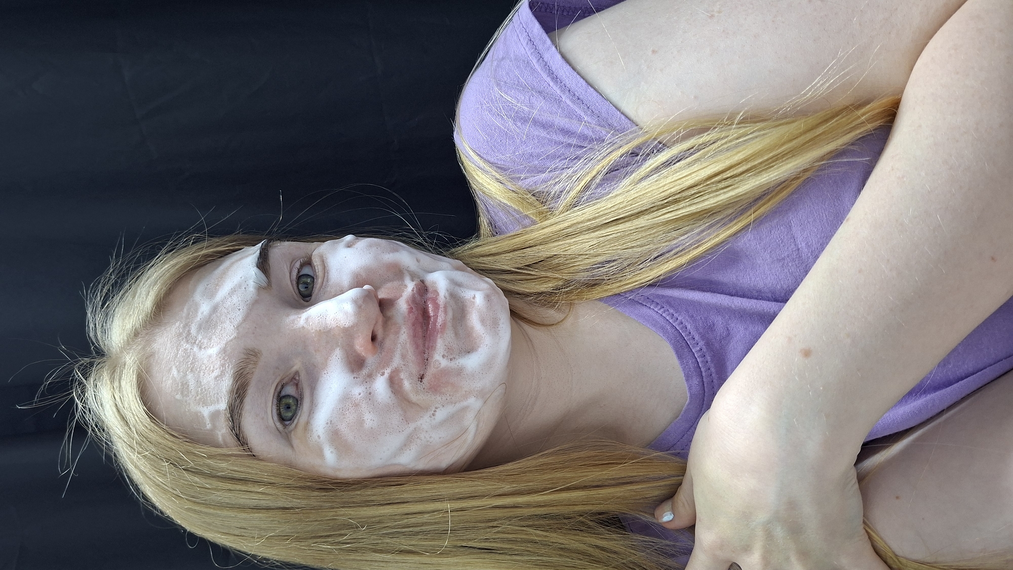 A fair-skinned woman with long blonde hair and green eyes lies on a black surface. Her face is covered in a white, foamy substance. She is wearing a light purple t-shirt. It is impossible to determine the exact location of this scene in Chaikovsky, Russia, from the image. The image only provides information about the subject and her immediate environment.
