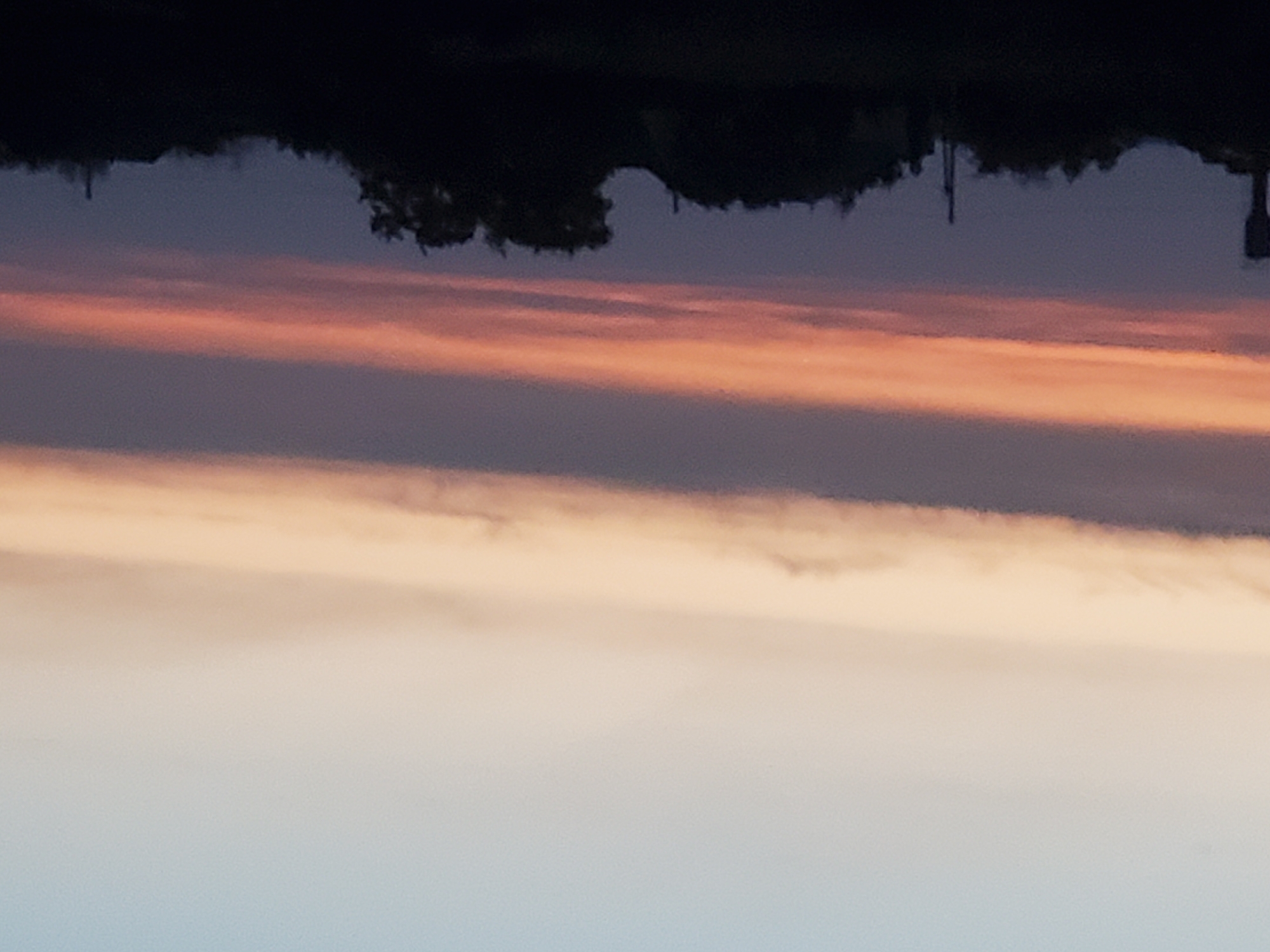 The image depicts a twilight sky over a silhouetted landscape. The sky transitions from a pale blue at the top to bands of peach and dark purple towards the horizon. A thin, vibrant orange band of clouds is prominent in the middle of the sky. The lower portion of the image is dominated by the dark silhouettes of trees and structures, including what appears to be a water tower on the left and possibly buildings or other man-made structures scattered throughout. The overall impression is of a rural or semi-rural setting at dusk. The location is specified as Konstantinovka, Moldova.