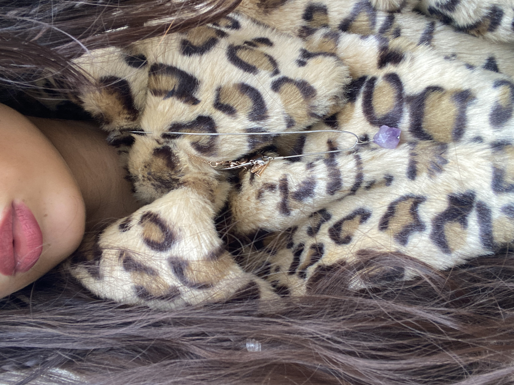 This close-up image features a person partially visible on the left side, showing their lower left cheek and lips. The lips are painted with a vibrant, muted pink or deep rose lipstick. Long, dark brown hair cascades around the frame, especially on the top, bottom, and left, intertwining with the main subject.
The central focus is a piece of leopard print fabric, likely a faux fur garment such as a coat or scarf. It displays a classic pattern of dark brown and black spots against a light beige background, characterized by its soft, plush texture.
A metallic, silver-toned decorative safety pin is fastened to the leopard print material. Attached to this pin are several small charms, including what appears to be a gold-colored initial or symbol and other metallic pendants. Towards the right end of the safety pin, a small, raw, faceted chunk of purple amethyst crystal is also secured, serving as a distinct accent.
The image is a tightly cropped, still shot, emphasizing the textures of the hair and fur, and the details of the accessories. No specific activity is taking place beyond the close-up presentation of these elements. There are no visible cues for time of day, weather, or discernible emotions from the partial view of the person's face. No text is visible within the image. The scene is situated in London, United Kingdom.