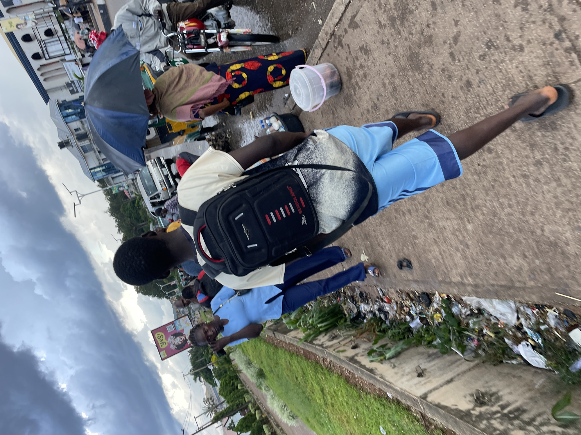 The image depicts a street scene in Barakin Lerawi, Nigeria, under a cloudy sky. In the foreground, a young Black male with dark skin, wearing a tan t-shirt and light blue shorts with dark blue stripes, walks away from the camera. He carries a black backpack with red accents. To his left, a Black female, wearing a light blue v-neck top and dark blue pants, walks while holding a black object to her ear, possibly a phone. She is walking next to a concrete drainage ditch filled with debris, plastic bags, and vegetation.
In the mid-ground, several people are visible. One individual, partially obscured, is under a blue and black umbrella. Behind them, a man on a motorcycle, wearing a gray jacket and dark pants, is stationary. Further back, a yellow and green three-wheeled vehicle is parked. A silver van is also visible on the road. A sign with the word "YES!" and images of faces is positioned on the left side of the road. Buildings with glass windows and architectural details are present in the background.
The environment appears to be a busy street with a sidewalk, a drainage channel, and traffic. The presence of trash in the ditch suggests potential sanitation issues. The overall atmosphere is that of an urban setting experiencing overcast weather.