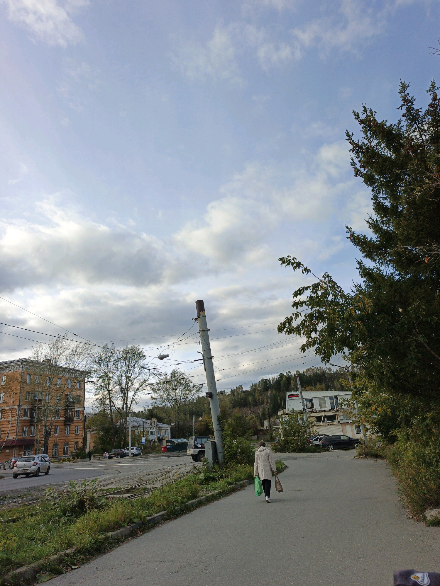 A lone figure walks away from the camera on a paved road, carrying two shopping bags. The person is wearing a light-colored coat and white shoes. The road curves to the left, leading towards an intersection with a large, orange apartment building on the left side of the image. Several cars are visible on the road, including a silver hatchback turning left and a dark SUV. Overhead power lines and a utility pole are present in the foreground and midground. In the background, there are trees and buildings, with a hillside covered in trees in the distance. The sky is partly cloudy with a mix of blue sky and gray clouds. This scene is captured on October 4, 2022, in Zlatoust, Russia.