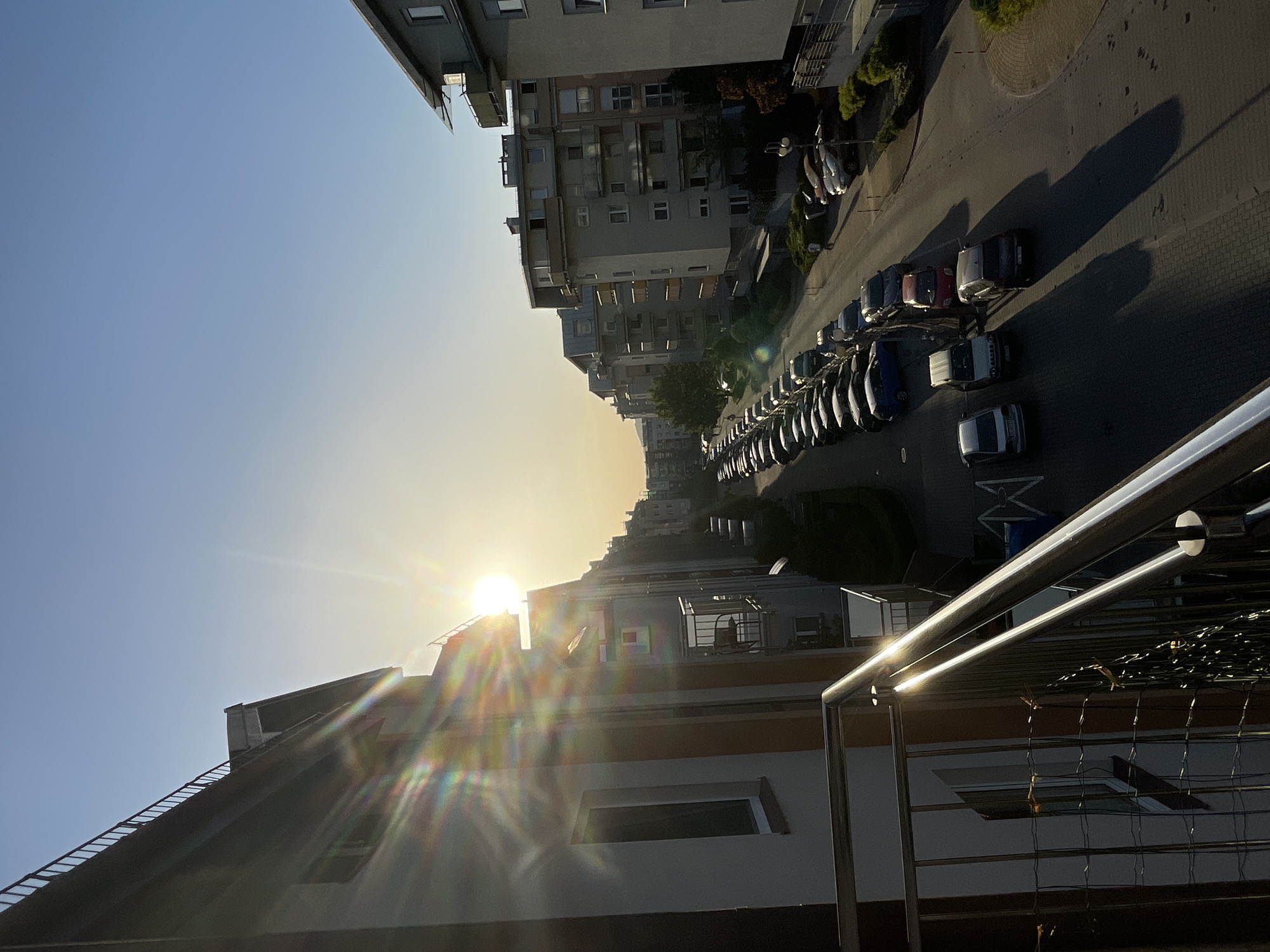 Here is a description of the image:
The image depicts an urban street scene at sunrise. The sun is low in the sky, causing lens flare. Buildings line the street, with cars parked along the side. A metal railing is visible in the foreground. The perspective suggests the photo was taken from a higher vantage point. The location appears to be residential, with multiple apartment buildings present.
