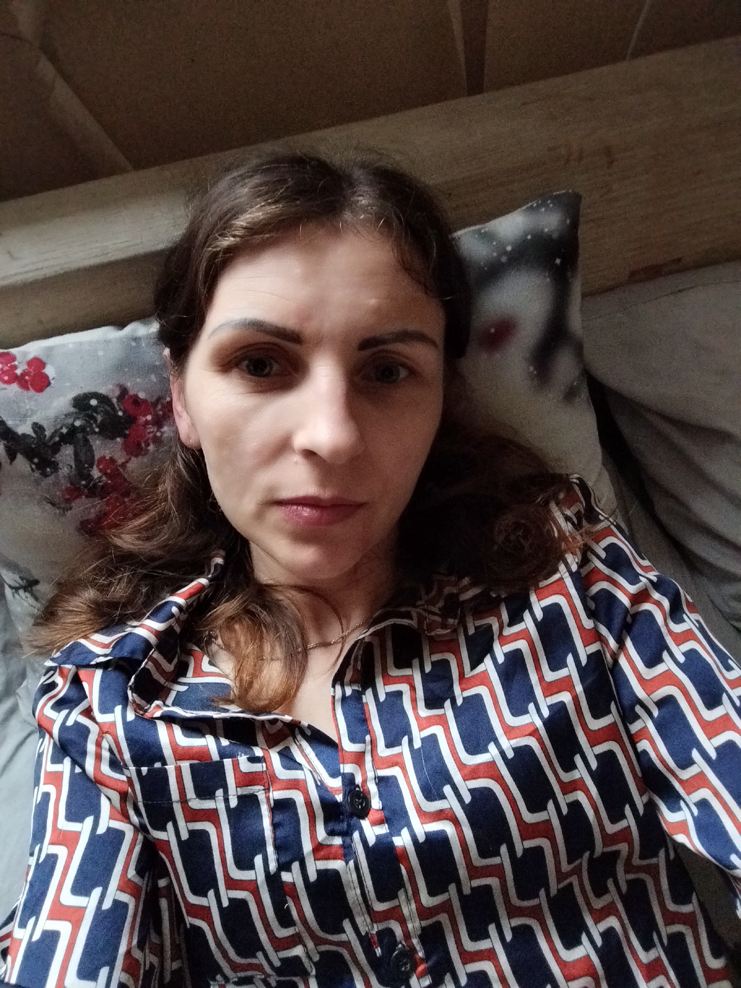 A woman with shoulder-length brown hair is lying down, taking a selfie. She is wearing a collared, button-up shirt with a geometric pattern of blue, red, and white shapes. She has on a delicate necklace. Behind her, a pillow with a floral pattern is visible, along with another pillow that appears to have a grey and white graphic. The background suggests an indoor setting, possibly a bedroom, with wooden beams visible above. The lighting is dim, and the image quality is moderate. The woman's facial expression is neutral.