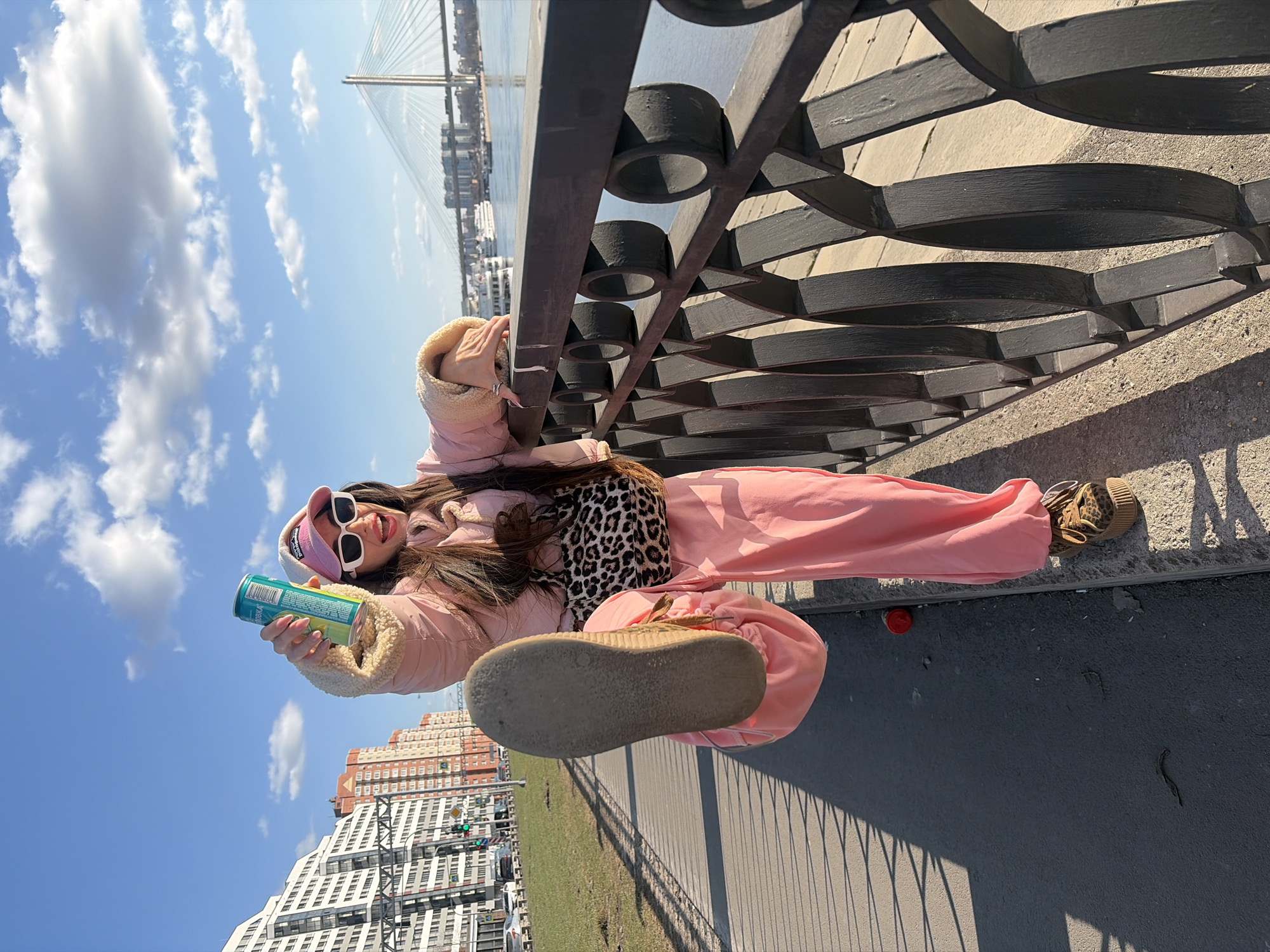 A young adult female is depicted in a dynamic pose on a paved embankment in Saint Petersburg, Russia, under a bright blue sky with scattered white clouds. The background features the cable-stayed bridge over the Korabelny Fairway, a section of the Western High-Speed Diameter (ZSD), spanning a wide body of water that appears to be the Neva River or its estuary. Large vessels are visible on the water beneath the bridge. To the left, several modern multi-story residential buildings line an urban street with visible traffic and streetlights.
The subject wears a pink puffy jacket with cream sherpa lining, pink baggy sweatpants, a pink baseball cap with a white Patagonia logo, white rectangular sunglasses, and brown leopard-print platform sneakers. A leopard-print fanny pack is slung across her torso. Her dark hair is long and falls over her shoulders. She exhibits an open-mouthed smile, looking directly at the camera. Her right leg is raised high, knee bent, with the worn sole of her platform sneaker facing the viewer. Her left hand rests on a dark, ornate metal railing bordering the embankment. In her raised right hand, she holds a teal-colored beverage can, displaying Russian text "ЧЕЛОВКА" (Chelovka) and a barcode. A red skateboard is partially visible on the ground near her left foot. The bright sunlight casts distinct shadows of the railing and the subject onto the pavement.