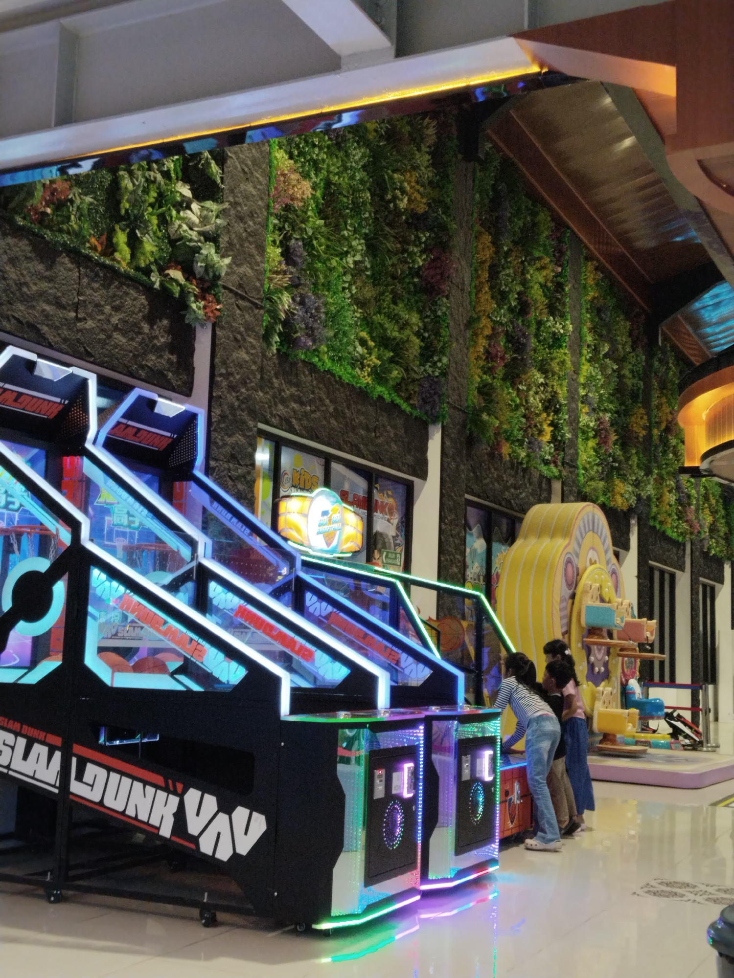 The image depicts an indoor arcade with a vertical garden wall and several arcade games. In the foreground, a "Slam Dunk" basketball arcade game is prominently displayed, featuring bright blue and white neon lights. To the right of the basketball game, three young females are interacting with other arcade machines. The environment appears to be a modern entertainment venue, possibly a mall or a dedicated amusement center. The vertical garden wall, composed of various green plants and flowers, adds a unique aesthetic to the background, contrasting with the artificial lights and structures of the arcade games. The overall scene suggests a lively and engaging atmosphere typical of an arcade. The specific location, Kecamatan Pati, Indonesia, is not visually evident from the image itself, but it is provided as context.