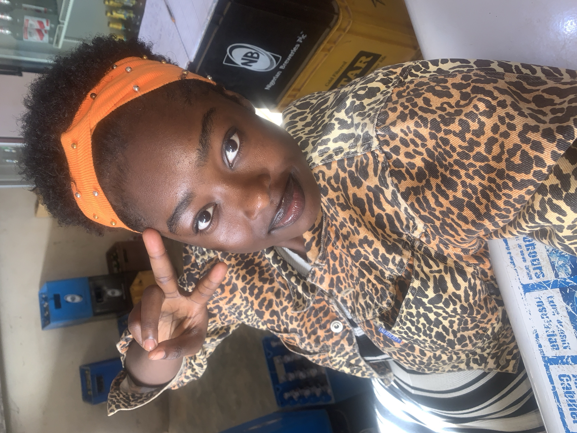The image shows a young Black woman with dark skin and curly hair, adorned with an orange beaded headband. She is wearing a leopard-print jacket over a horizontally striped shirt. Her right hand is raised, fingers spread in a peace sign. She is indoors, with stacks of blue and yellow crates visible in the background. One crate has the "NB Nigerian Breweries Plc" logo. The environment suggests a retail or storage area, possibly a bar or shop. The lighting is bright, indicating it is daytime. The setting is consistent with locations found in Jos, Nigeria.