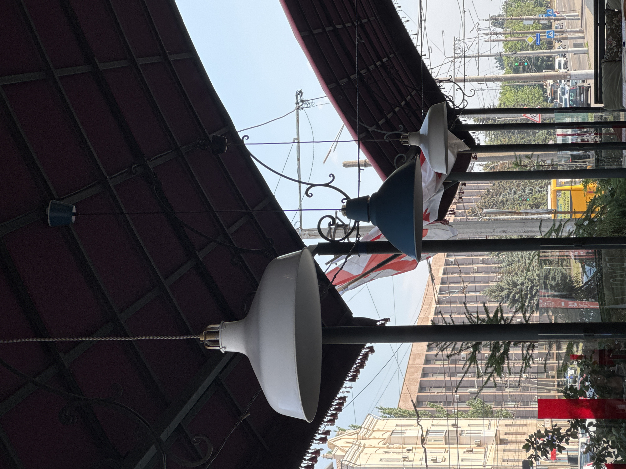 The image shows an outdoor seating area, likely a cafe or restaurant, with a red awning overhead. Two large, upside-down pendant lamps hang from wires. One lamp is white and bell-shaped, while the other is blue and has a wider, flatter shade. Both lamps appear to be made of metal. Decorative wrought iron scrollwork is visible along the underside of the awning.
Beyond the seating area, the scene opens up to an urban streetscape. A light pole with numerous wires stretches across the frame, indicating an area with public transportation, possibly trams or trolleybuses, which are common in Ukrainian cities. Trees line the street, and several buildings of varying heights are visible in the background, including a multi-story apartment building with many windows and air conditioning units. Traffic, including cars and possibly a yellow bus, can be seen on the street, along with traffic signals and road signs. The sky is clear and bright, suggesting it is daytime, likely sunny. The image has a somewhat distorted perspective, possibly due to the camera angle or a wide-angle lens. There is some text on the yellow bus, but it is not legible. The overall impression is a bustling urban environment on a bright day.