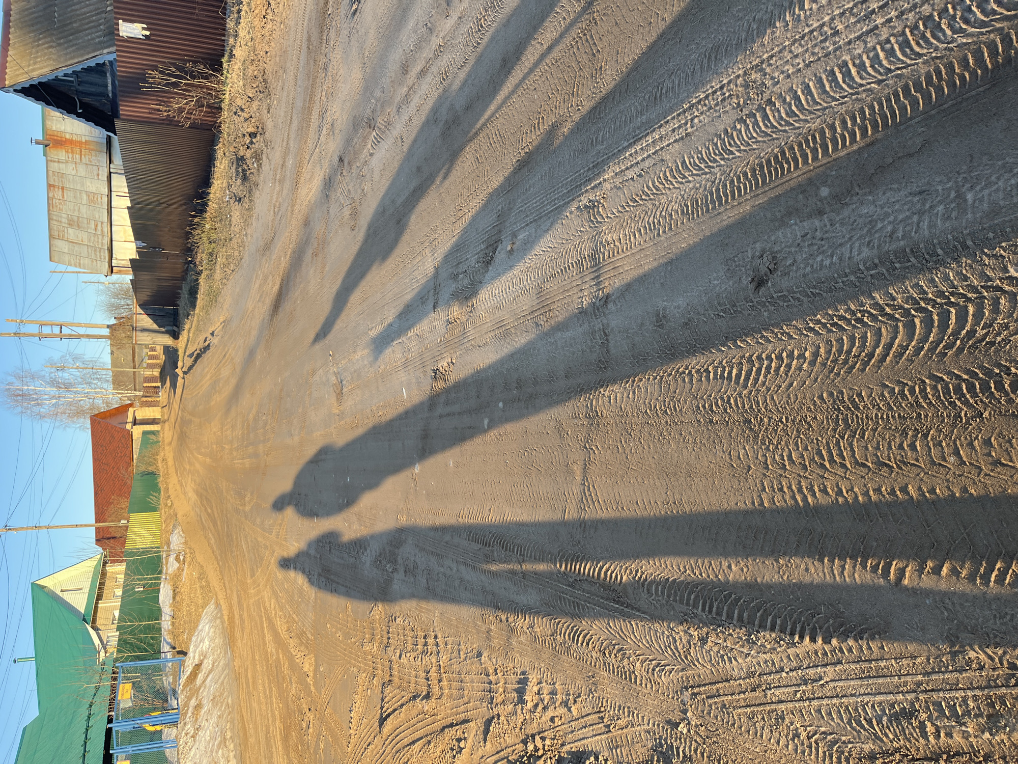 The image depicts a dirt road with tire tracks in Chaikovsky, Russia. Shadows of trees or buildings cast long stripes across the road. The road is flanked by buildings, including a metal-clad structure on the left and houses with varying roof colors (green, red) on the right. Utility poles and wires are visible in the upper left corner. Two darker shadows resembling human figures are cast on the road further away. The sky is light blue.