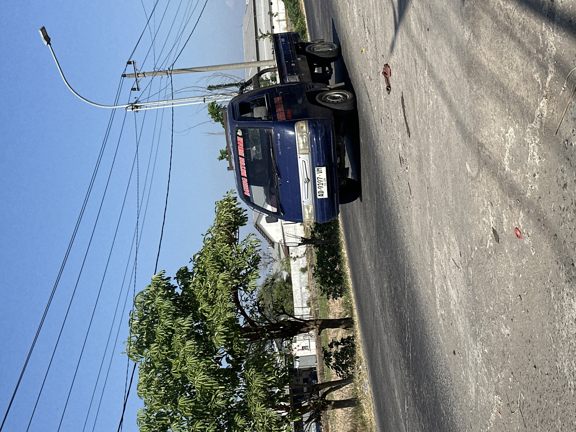 A blue Suzuki Carry pickup truck drives down a paved road in Parangjoro, Indonesia. The truck has a red banner across the top of the windshield with text that appears to read "AGUNG BETON INDAH". The license plate reads "AD 9197 VM". The truck is facing left, and the image is taken from the side. A large tree with green leaves is on the left side of the road, and a utility pole with wires and a street lamp is on the right. In the background, there are buildings and fields. There are also some red flip-flops lying on the side of the road. The sky is clear and blue.