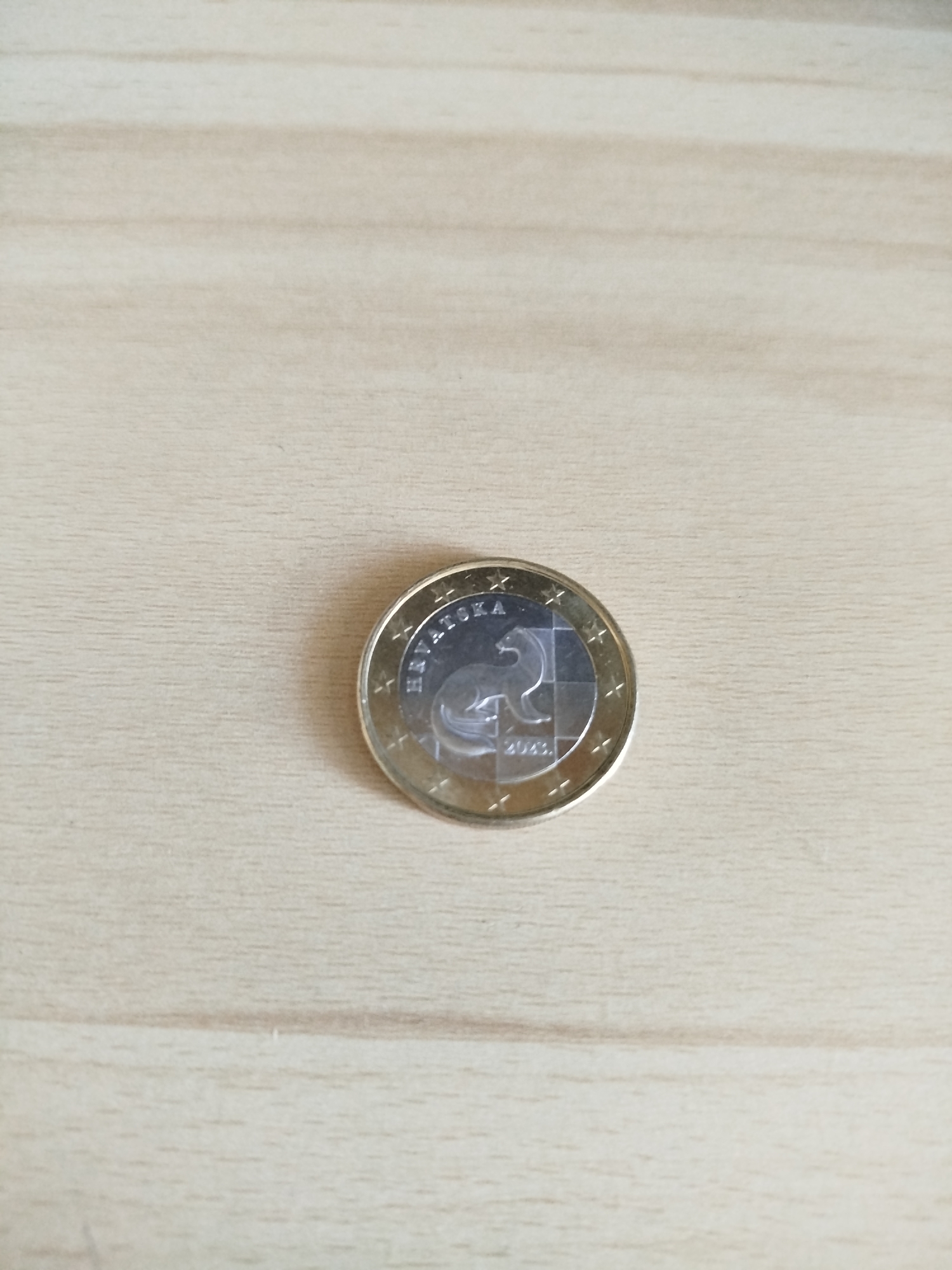 A single euro coin from Croatia is centered on a light beige, wooden surface. The coin features a two-tone design with a silver inner circle and a gold outer ring. The word "HRVATSKA" is visible along the upper edge of the coin, and the year "2023" is printed below the central image. The scene is minimalist, with the coin as the sole subject, set against the plain backdrop of the wooden surface.