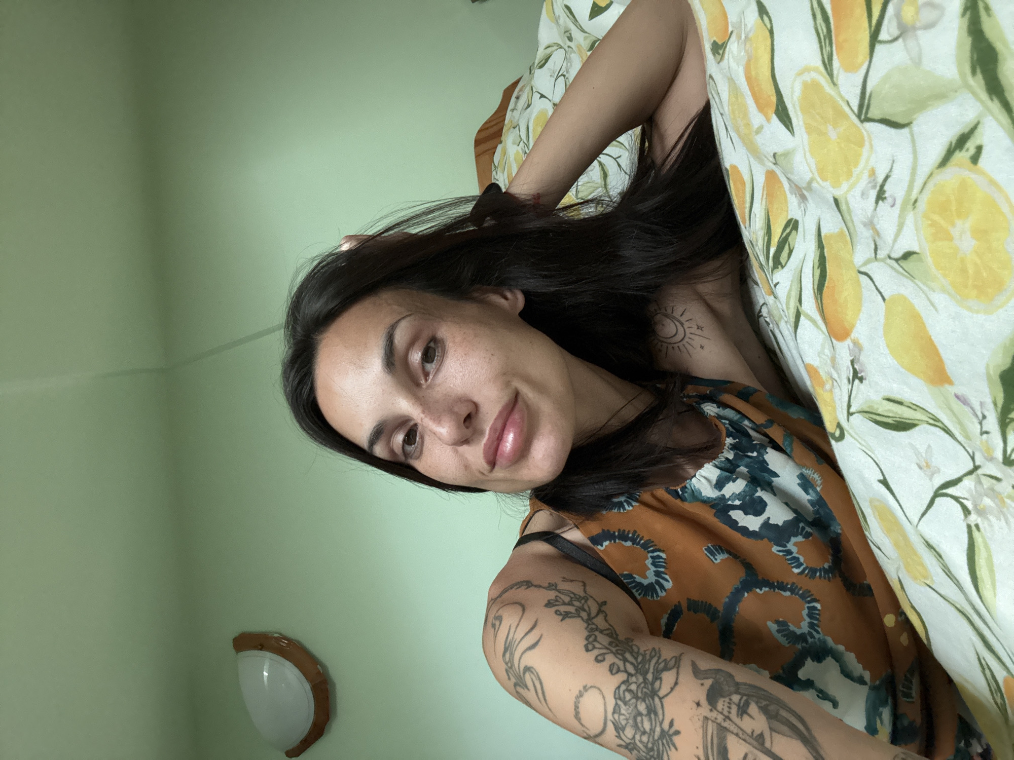 A woman with dark hair and light skin is in a bed with a lemon-patterned sheet. She has tattoos on her right arm and collarbone. The background wall is light green, with a wall sconce visible. The location is Pleven, Bulgaria.