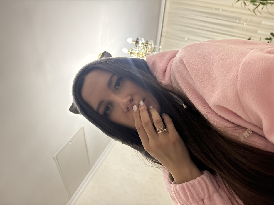This indoor image, located in Vysokinichi, Russia, features an adult female subject with long, dark hair and light-colored eyes. She is wearing a grey hooded garment with two pointed, cat-like ears on the hood, over a pink fleece hoodie. Her right hand, adorned with four thin rings on the ring finger and white nail polish, is positioned with fingers resting near her mouth. The subject is looking directly forward. The background reveals a white ceiling with a light-colored access panel on the upper left, and a multi-lamp gold-toned chandelier with crystal elements on the upper right. A light-colored curtain is partially visible behind the chandelier, and a faint outline of a plant is discernible in the lower right corner. The walls are light-toned with white crown molding at the ceiling line.