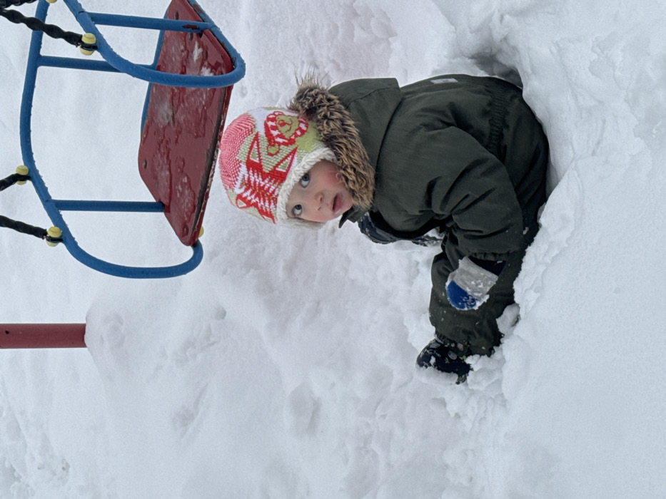 The image shows a young child playing in deep snow, captured from a slightly elevated perspective. The child is positioned in what appears to be a snowdrift or a small snow tunnel, with a significant amount of snow surrounding them, reaching up to their torso. The child is looking directly at the camera with a curious and slightly wide-eyed expression, their tongue slightly out.
The child is warmly dressed in winter attire, featuring a multi-colored knitted winter hat with a pom-pom and a light-colored fur trim. The hat has patterns in pink, white, and red, including what looks like stylized animal faces or festive designs. They are wearing a dark green, possibly olive green, padded winter jacket with a fur-lined hood. One blue and white mitten is visible on their right hand, which appears to be resting on their knee or in the snow. Their lower body and legs are mostly submerged in the snow, but a dark winter boot is partially visible on their right foot.
In the background and to the left, part of a blue and red playground swing set is visible. The blue metal frame of the swing structure is prominent, along with a red swing seat and yellow connection points for the swing chains. A red support beam is also seen at the bottom left. The entire scene is covered in pristine white snow, indicating a cold, snowy day. The lighting is soft and even, suggesting an overcast sky. The image itself is rotated 90 degrees clockwise, making the child and playground equipment appear on their sides.
