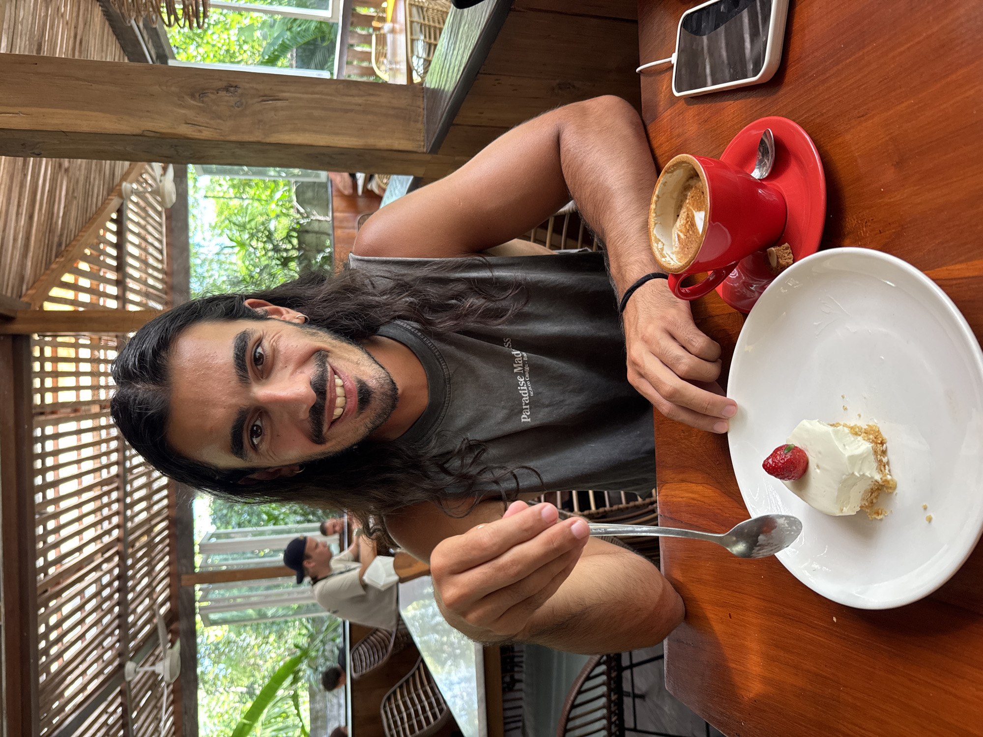 A man with long dark hair is sitting at a wooden table indoors. He is smiling and holding a spoon towards the camera. On the table is a white plate with a piece of cheesecake topped with a strawberry. There is also an empty red coffee cup and a mobile phone. In the background, through a window, are other people and green plants. The location is Kuta Selatan, Indonesia.