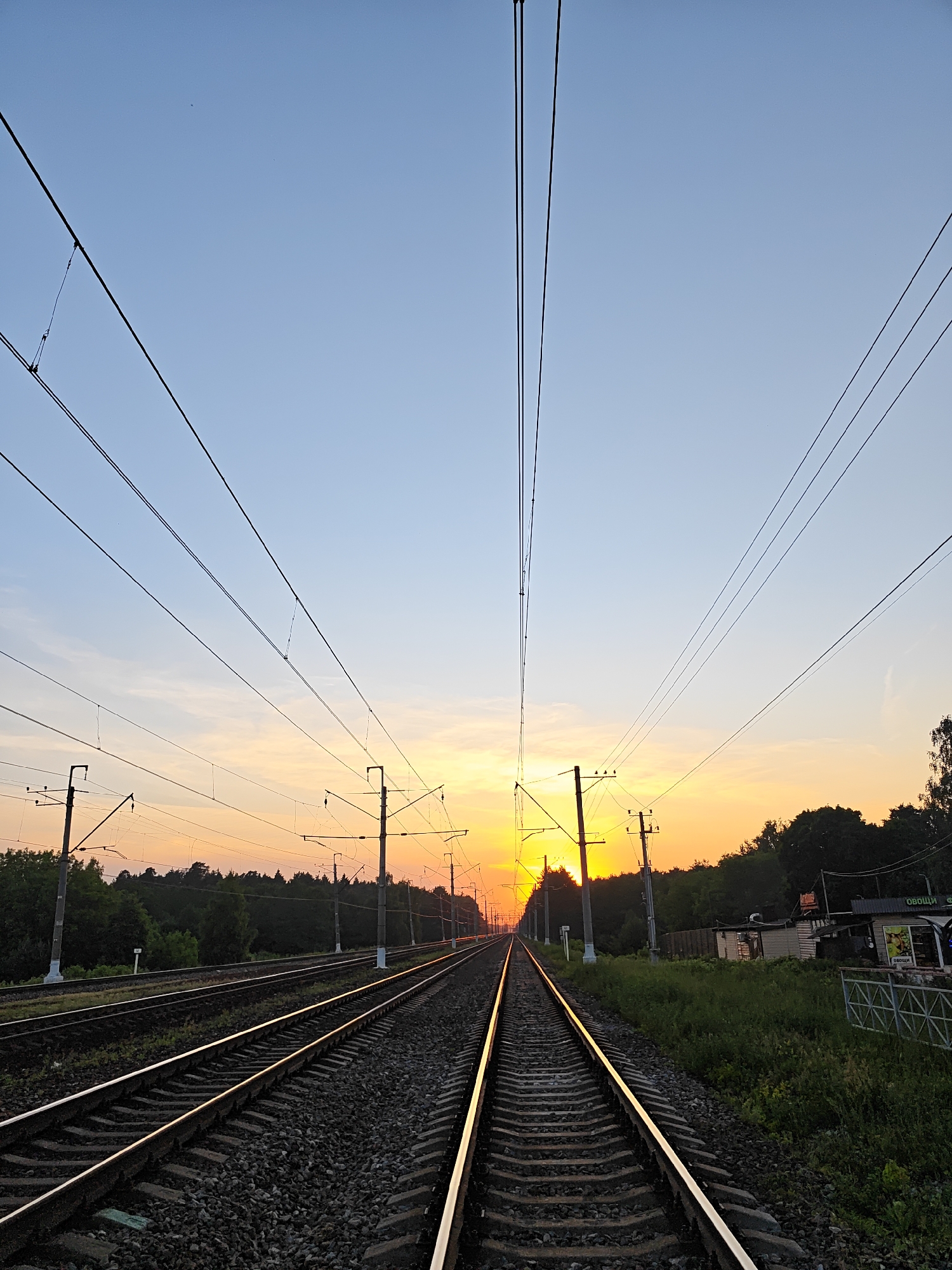 The image depicts a sunset over multiple parallel railroad tracks in Kratovo, Russia. The sky transitions from a light blue at the zenith to vibrant orange and yellow hues at the horizon, where the sun is low, creating a silhouetted effect. Several utility poles, supporting an intricate network of overhead electrical catenary lines, extend along both sides of the tracks, converging towards the distant sunset point. The steel rails brightly reflect the warm light of the setting sun, particularly on the inner rails of the two central tracks. Ballast stone and wooden sleepers form the track beds. Dense green vegetation, appearing as dark silhouettes, borders the tracks, forming a distant treeline. In the mid-ground on the right, alongside the tracks, are small, simple structures, one of which features a sign displaying the Russian word "ОВОЩИ" (Vegetables), identifying it as a produce stand. A metal fence is visible in front of these structures. No individuals are present in the scene.