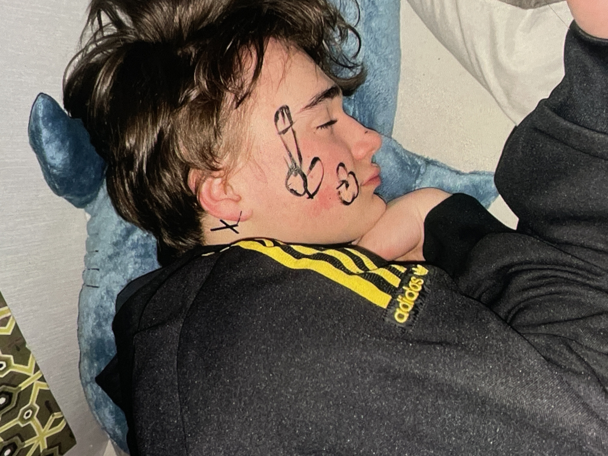A person with dark hair is sleeping on their side, facing right. They are wearing a black Adidas tracksuit with yellow stripes. There are drawings on their face, including what appears to be a penis and a smiley face. The person is lying on a blue pillow and a white pillow. The background is out of focus and shows a patterned surface and a wall. The image does not provide any information about the city of Harmanli, Bulgaria.