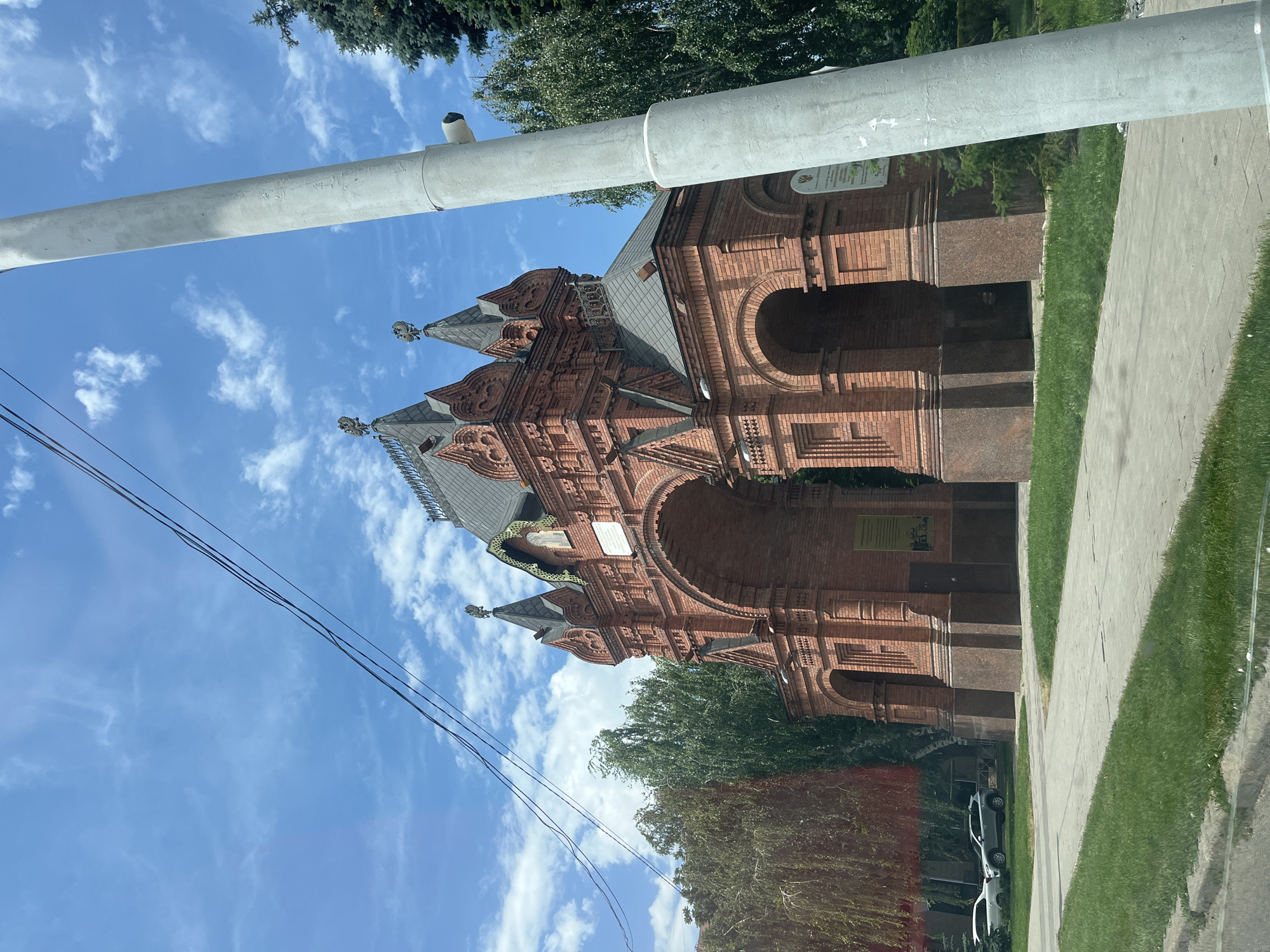 An ornate red brick archway stands prominently in the image, with intricate architectural details and multiple turrets topped with double-headed eagles. The structure appears to be made of red brick with dark stone bases. To the right of the archway, a tall, light gray cylindrical pole with a security camera attached leans into the frame. In the background, lush green trees and a bright blue sky with scattered white clouds complete the scene. In the lower left corner, a glimpse of cars and a paved walkway suggest an urban setting. The archway is identified as the Glory Arch in Krasnodar, Russia, a monument commemorating military victories.