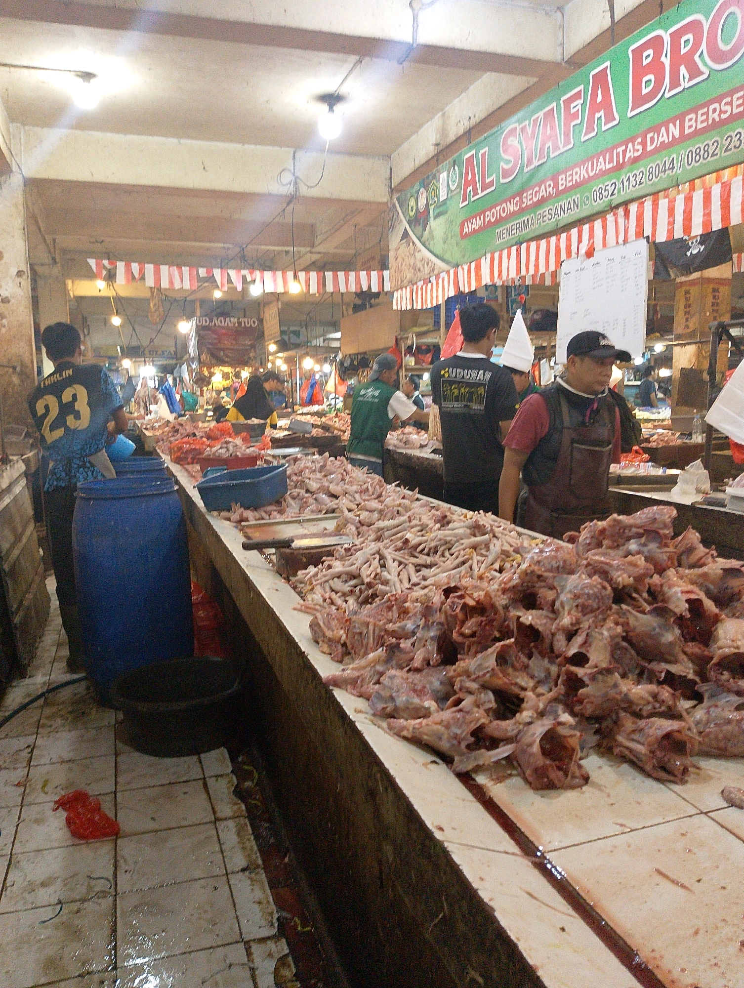The image depicts a bustling marketplace scene in Lembang, Indonesia, likely a section dedicated to the sale of poultry. The foreground is dominated by a long counter laden with an extensive display of raw chicken parts, including carcasses and feet, arranged in piles. Several workers, dressed in practical attire, are present. One individual in a blue shirt with the number '23' on the back stands near a large blue barrel. Another worker, wearing a black cap and a dark apron over a maroon shirt, appears to be attending to the meat. In the background, stalls with various goods are visible, along with hanging decorations and signage. The overall atmosphere suggests an active commercial environment, with the focus on fresh produce.